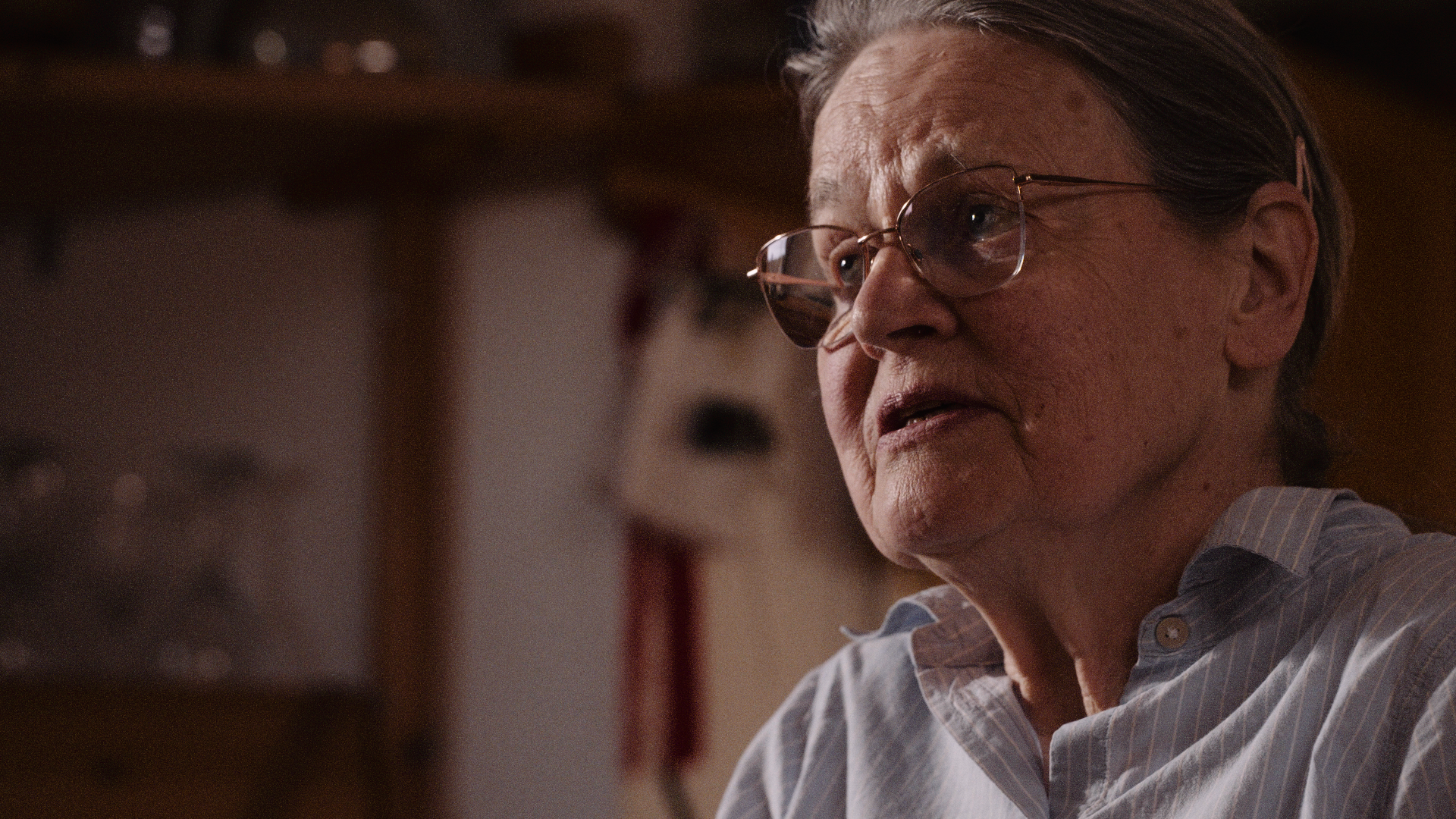 Close-up of an elderly woman with glasses and gray hair, wearing a striped shirt, sitting and talking in a cozy indoor setting.