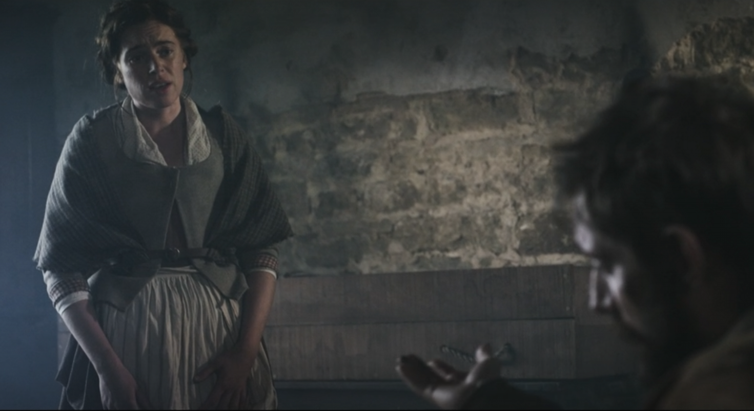 A woman with dark hair, wearing a gray scarf and a checkered shawl, standing and speaking to a man seated, who is gesturing with his hand, in a dimly lit rustic room with exposed stone walls.