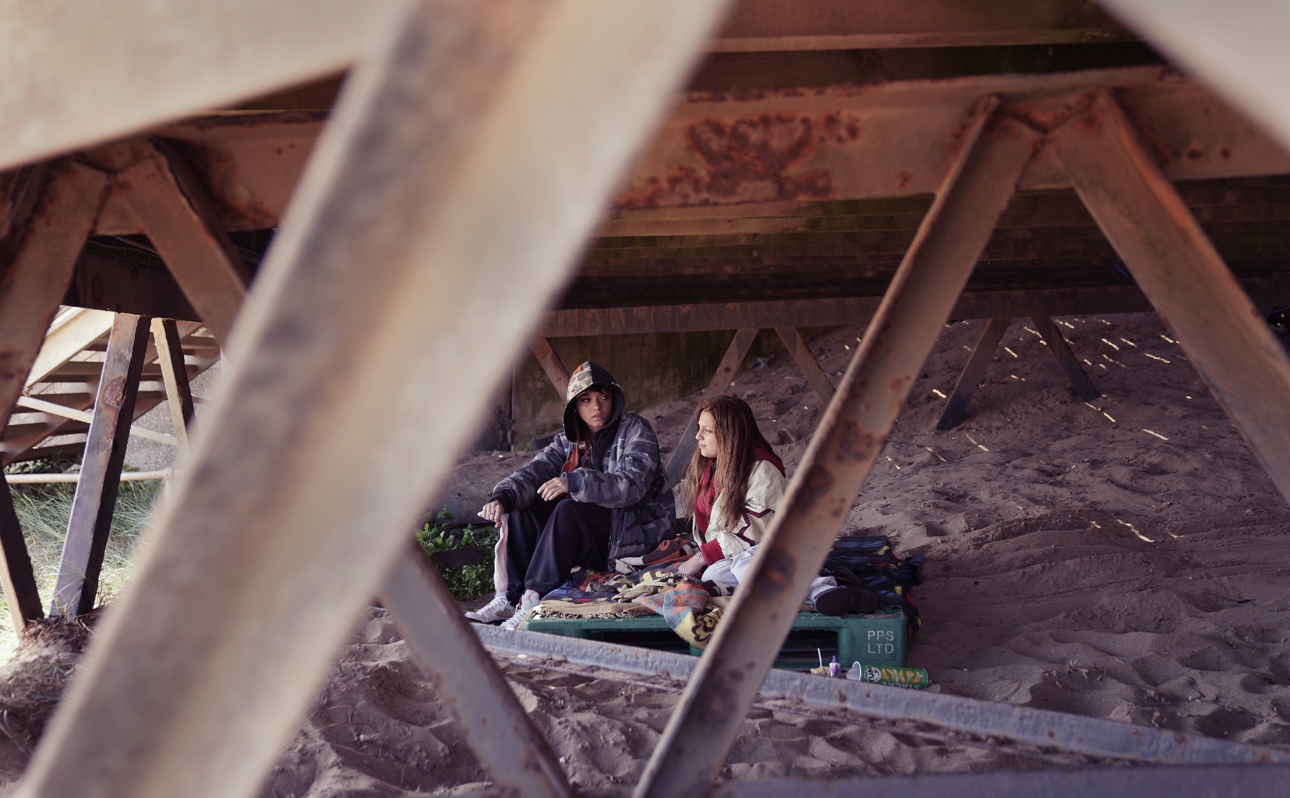 Two women sitting on blankets under a wooden pier on a sandy beach, seen through the pier's lattice structure.