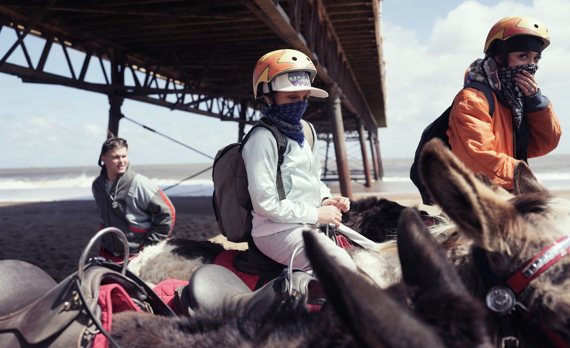 Three children with face masks and helmets riding donkeys on a beach, with a pier structure in the background under a partly cloudy sky.