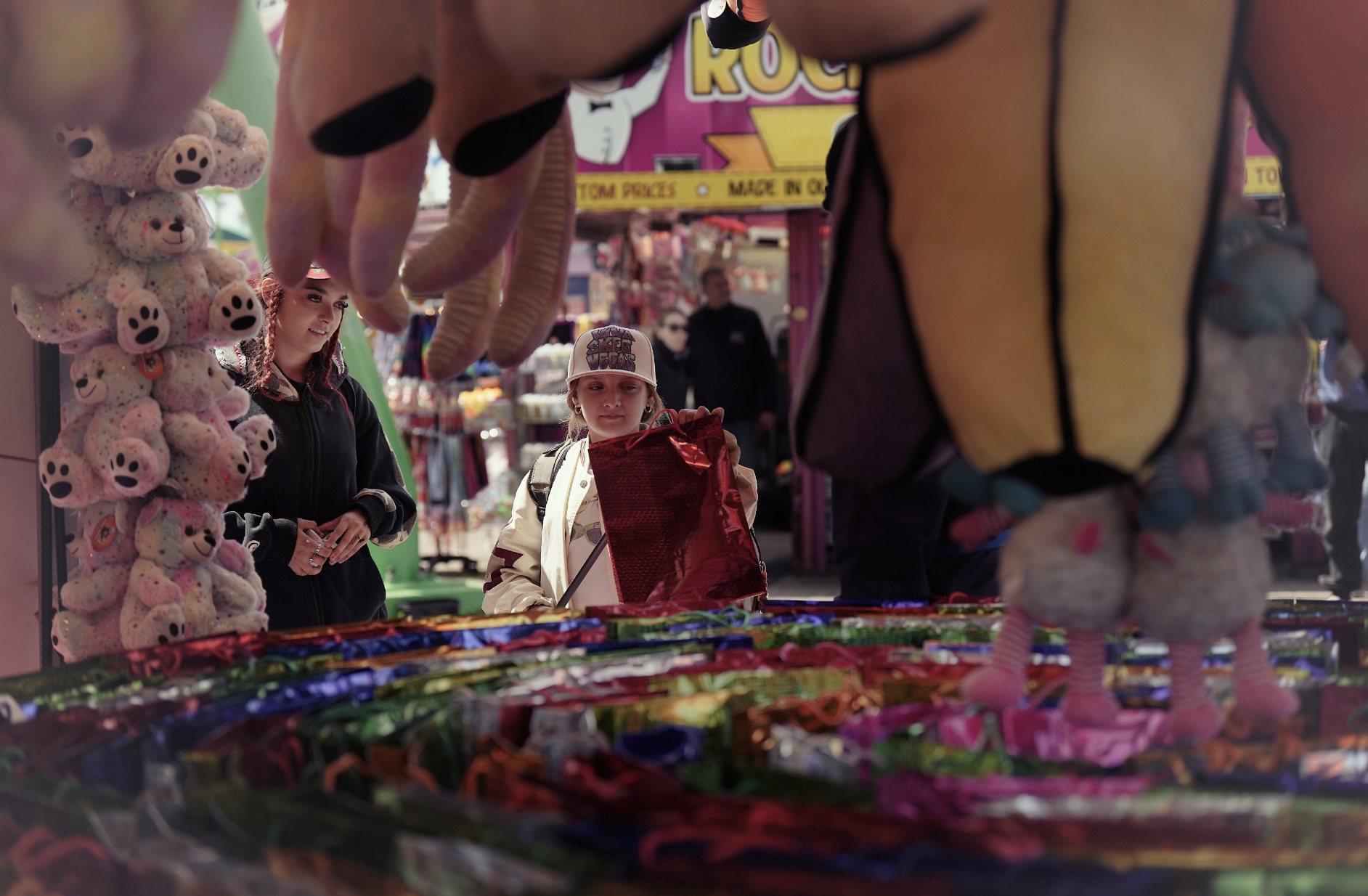 View of two young women at a market stall filled with colorful candies, with plush toys hanging in the foreground.