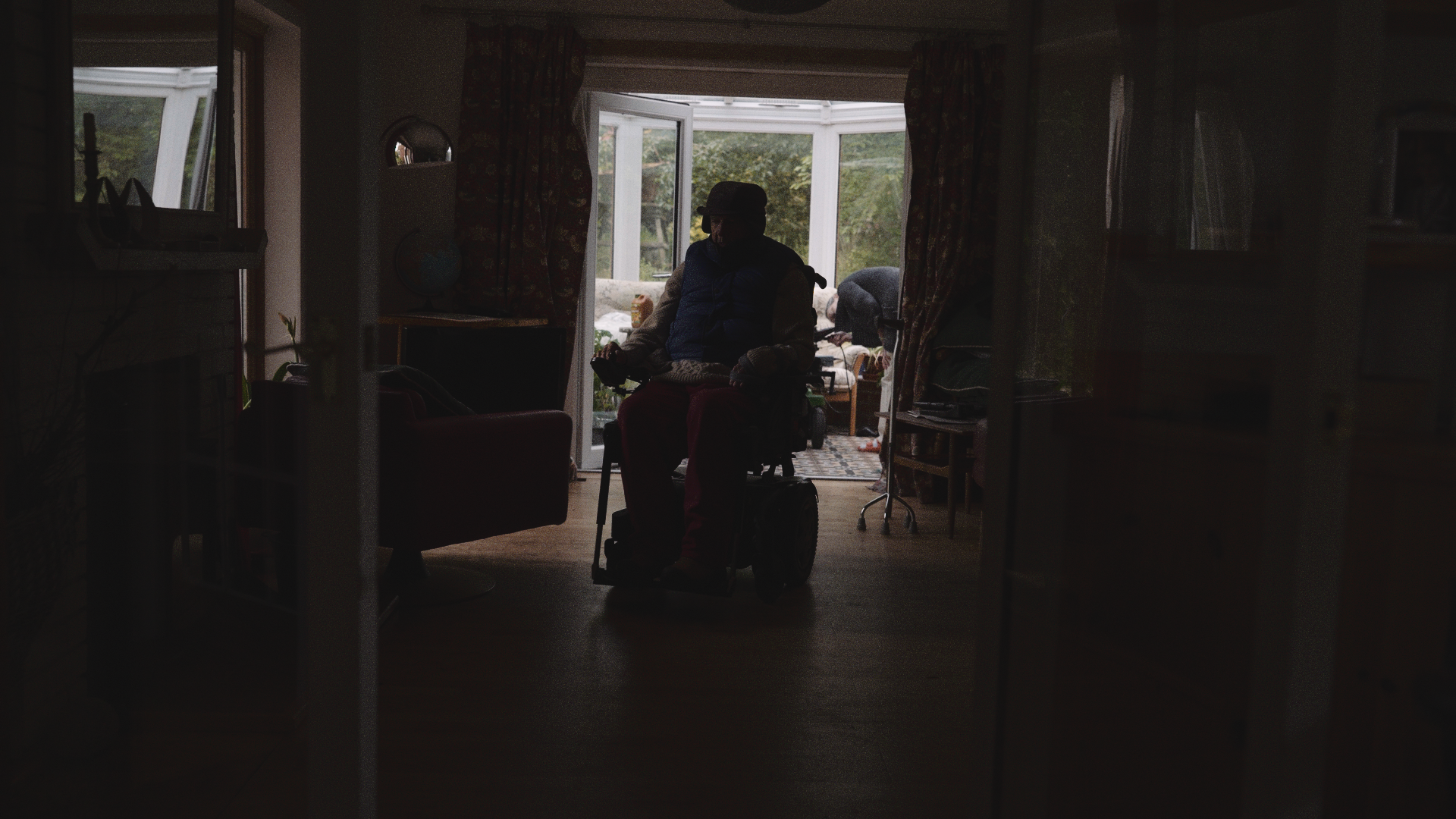 Silhouette of a person in a wheelchair inside a dimly lit room, with open glass doors leading to a sunroom with bright natural light outside.