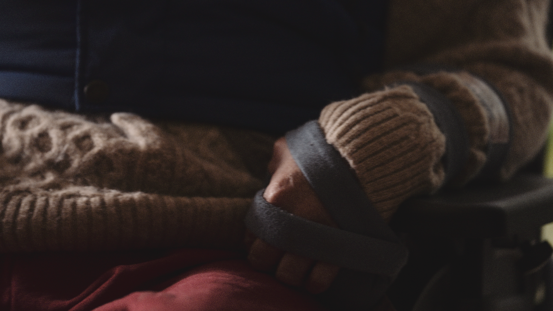 Close-up of a person's hand holding a smartphone, wearing a brown ribbed sweater and dark jacket, with a textured brown blanket nearby.
