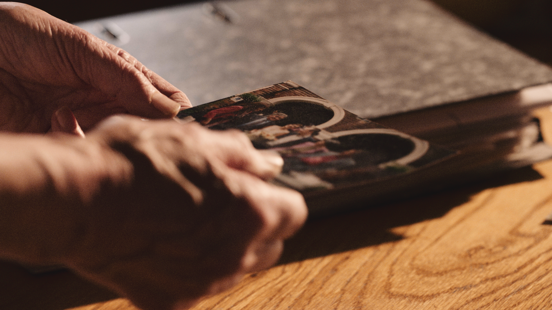Close-up of elderly person's hand holding a photo album or scrapbook on a wooden table.