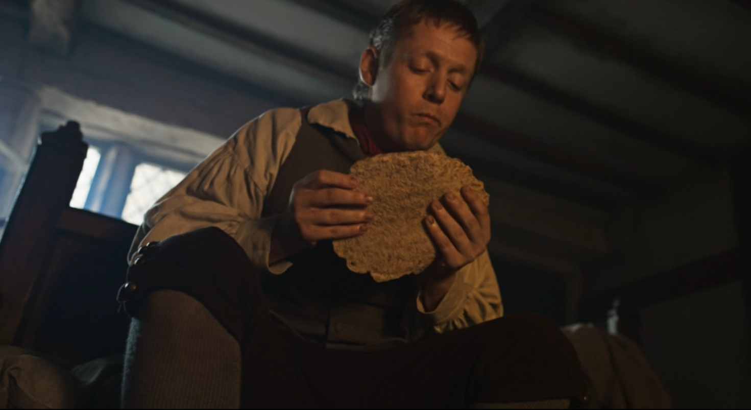 A man sitting on a bed indoors, holding and eating a large piece of bread or biscuit.
