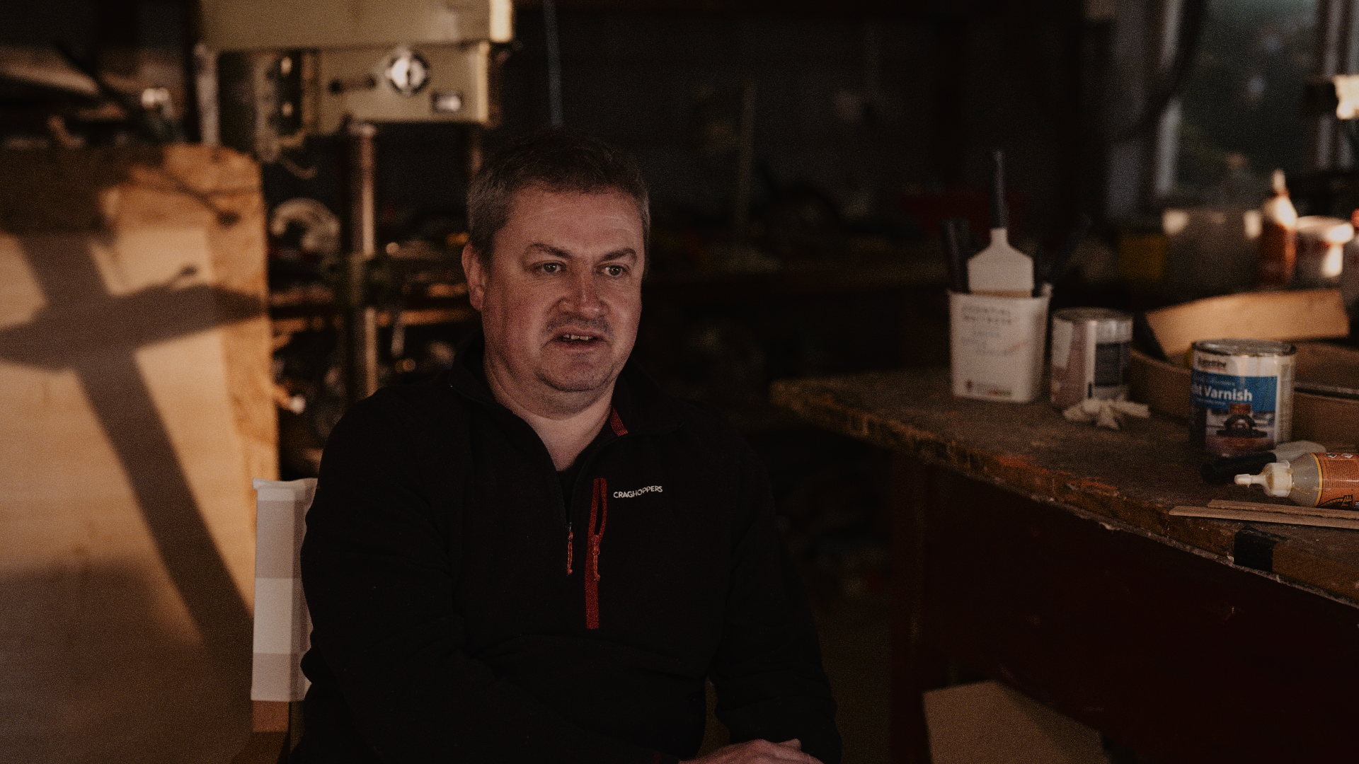 A man with short dark hair and light skin sitting at a cluttered workbench in a workshop, wearing a black jacket with red accents. There are various tools, paint cans, and supplies on the workbench behind him.