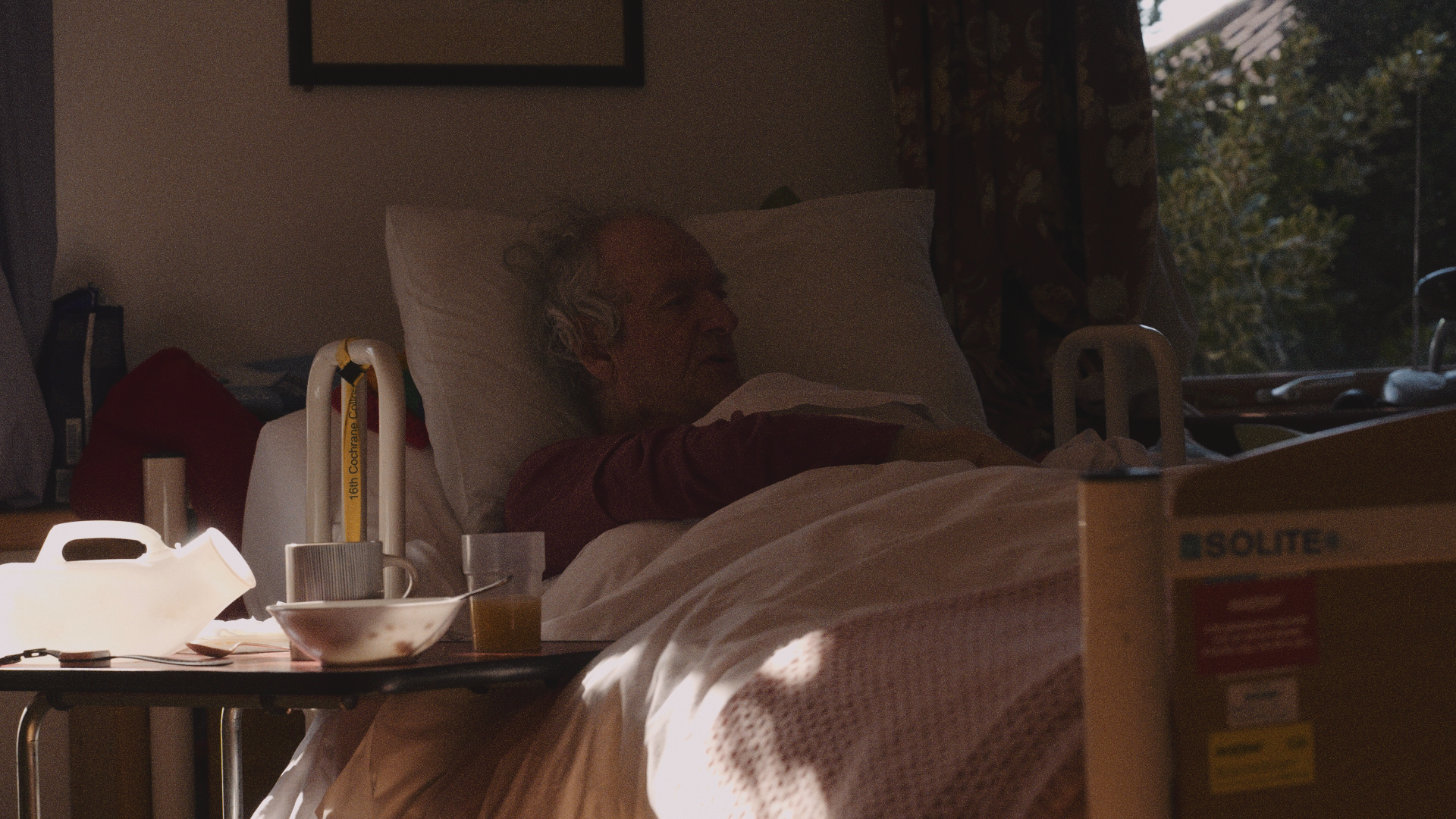An elderly man with gray hair resting in a hospital bed, illuminated by soft natural light from a window with floral curtains, surrounded by hospital equipment and personal items.