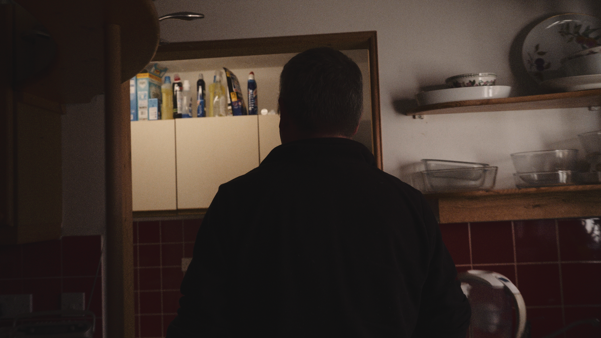 A person with short dark hair viewed from behind, standing in a kitchen with shelves holding bowls and a mirror reflecting cabinet with cleaning supplies.