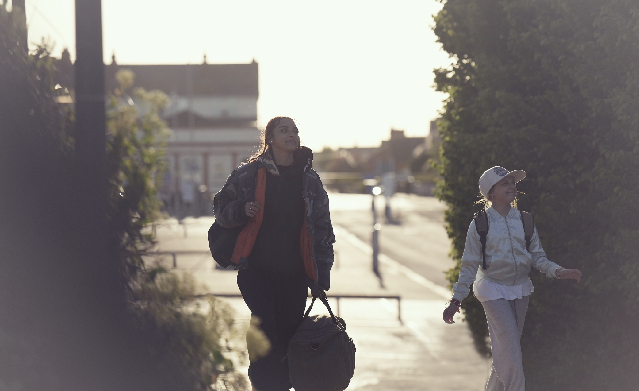 Two young girls walking outdoors during the daytime, one wearing a camouflage jacket and the other a white jacket and cap, carrying backpacks, with sunlight creating a backlit scene.