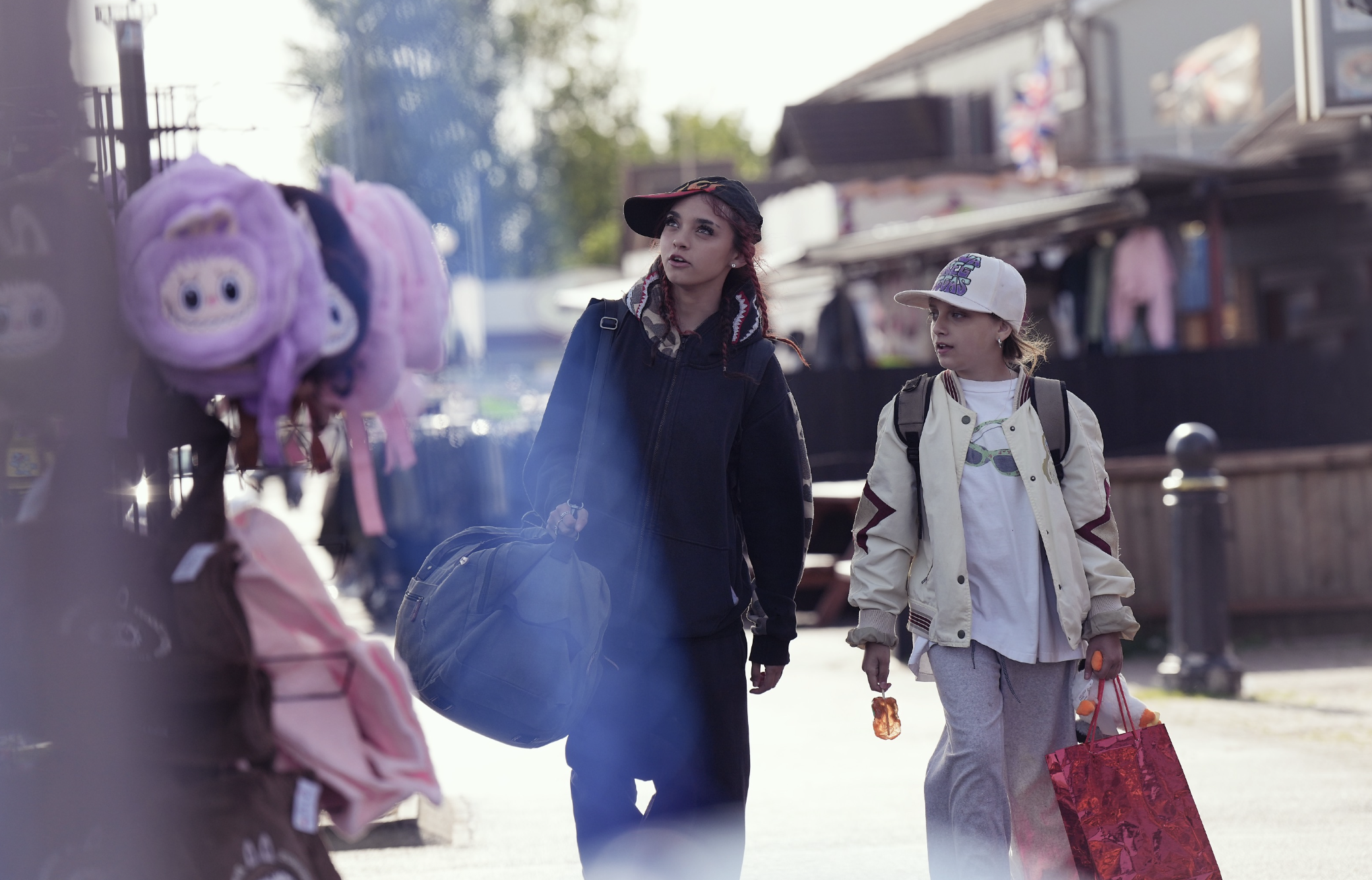 Two young girls walking outdoors near a store with plush toys on display, one wearing a black cap and jacket, the other with a baseball cap and light jacket, both carrying shopping bags.