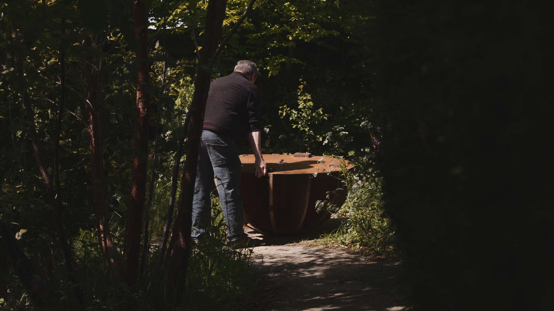 A man with gray hair wearing a black jacket and jeans is outdoors, bending over and working on constructing or repairing a wooden structure, possibly a large round table, in a wooded area with dense green foliage.