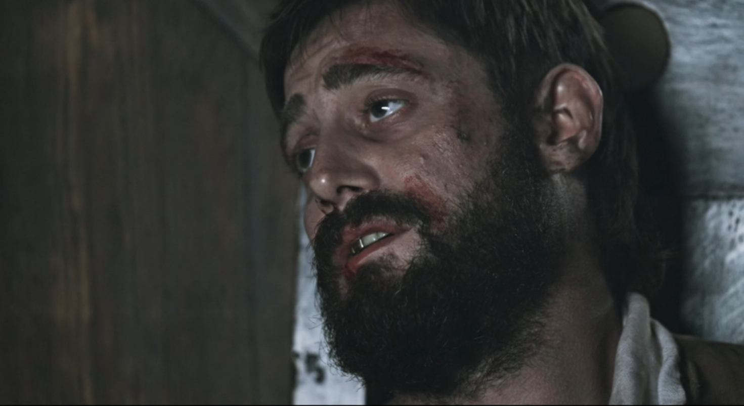 A close-up of a man with a beard, injuries, and blood on his face lying on a wooden surface.