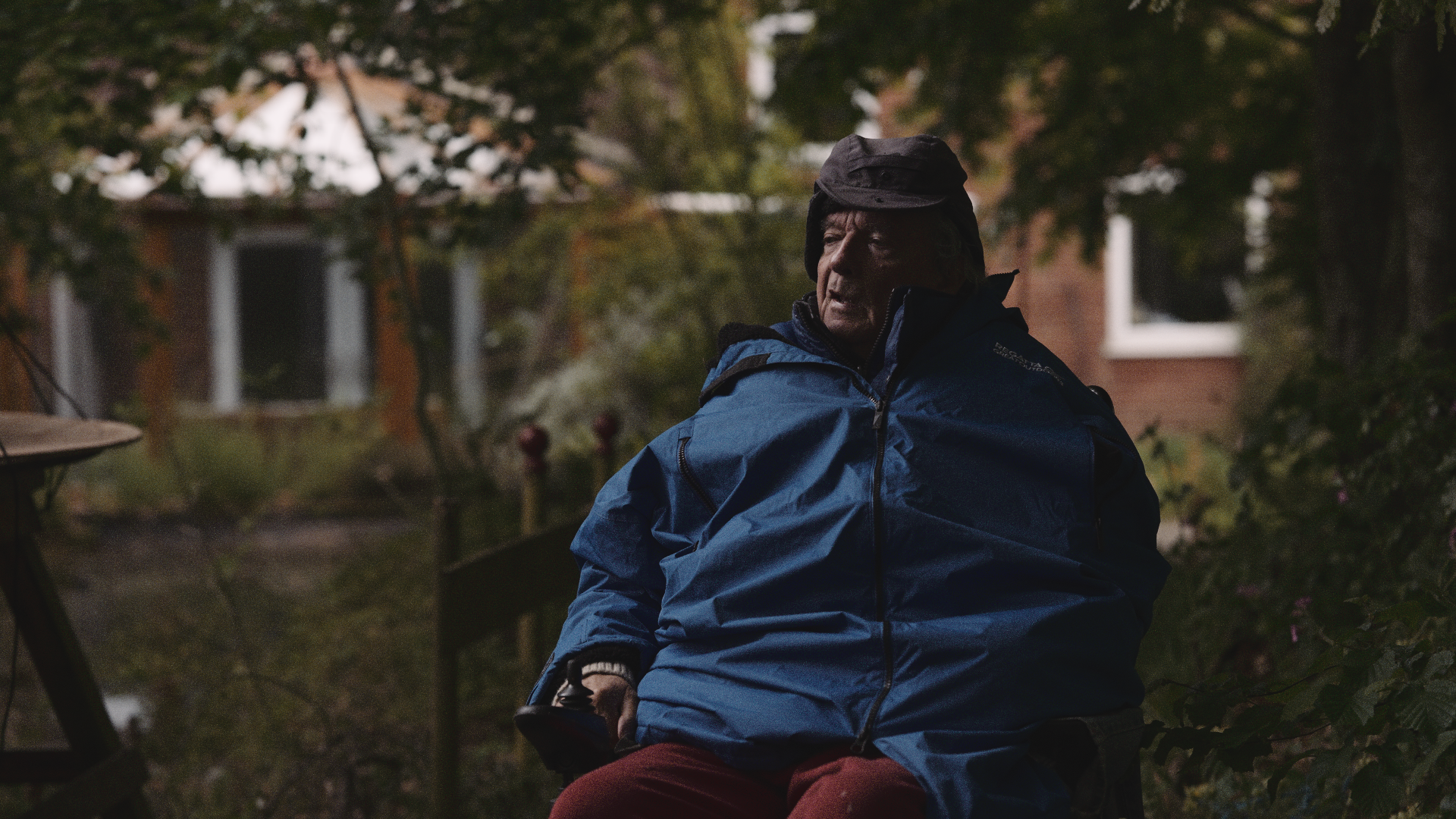 An older man sitting outdoors in a blue jacket and black hat, with trees and a brick house in the background.