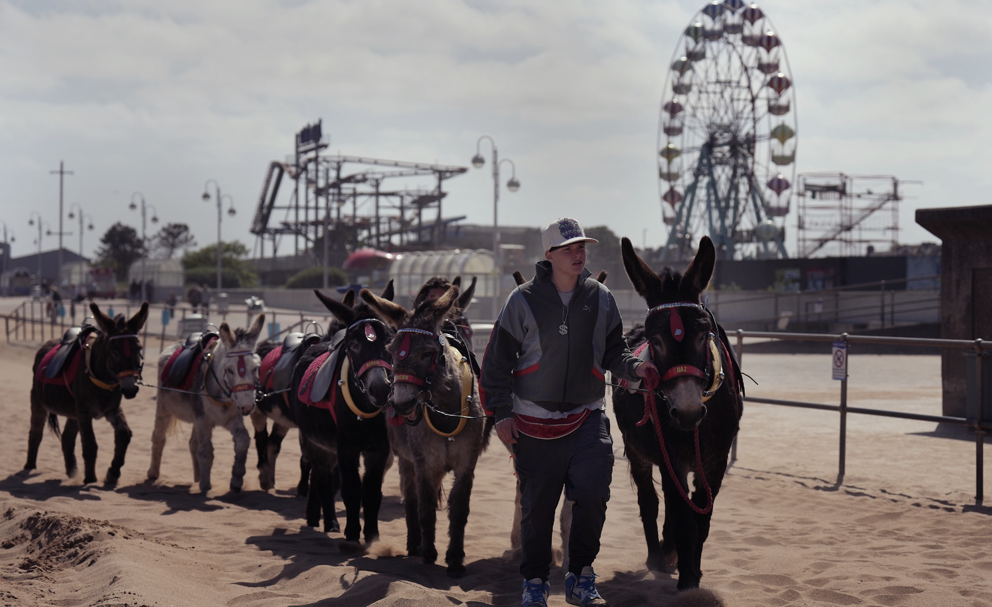 A young man guiding a line of small donkeys on a sandy fairground. In the background, there is a large Ferris wheel and amusement park rides under a cloudy sky.