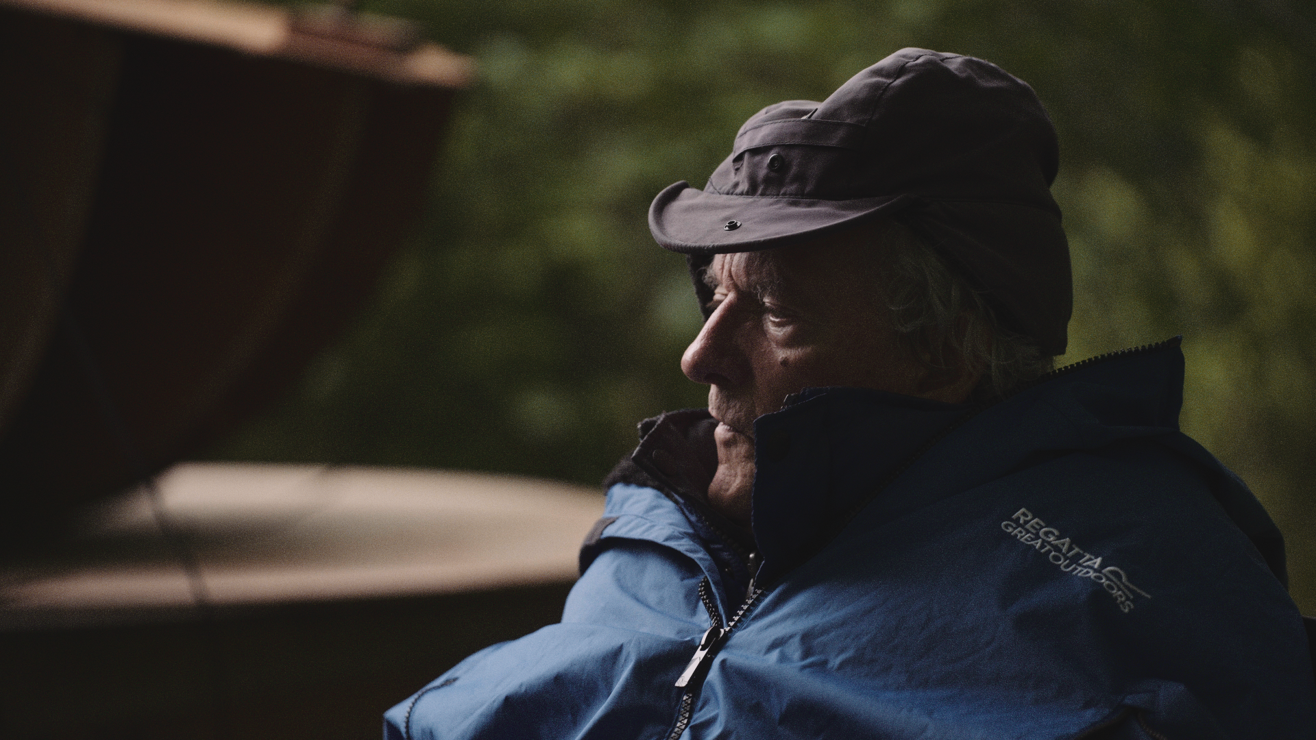 A man wearing a black bucket hat and a blue outdoor jacket, sitting outdoors and looking towards the left with a serious expression.