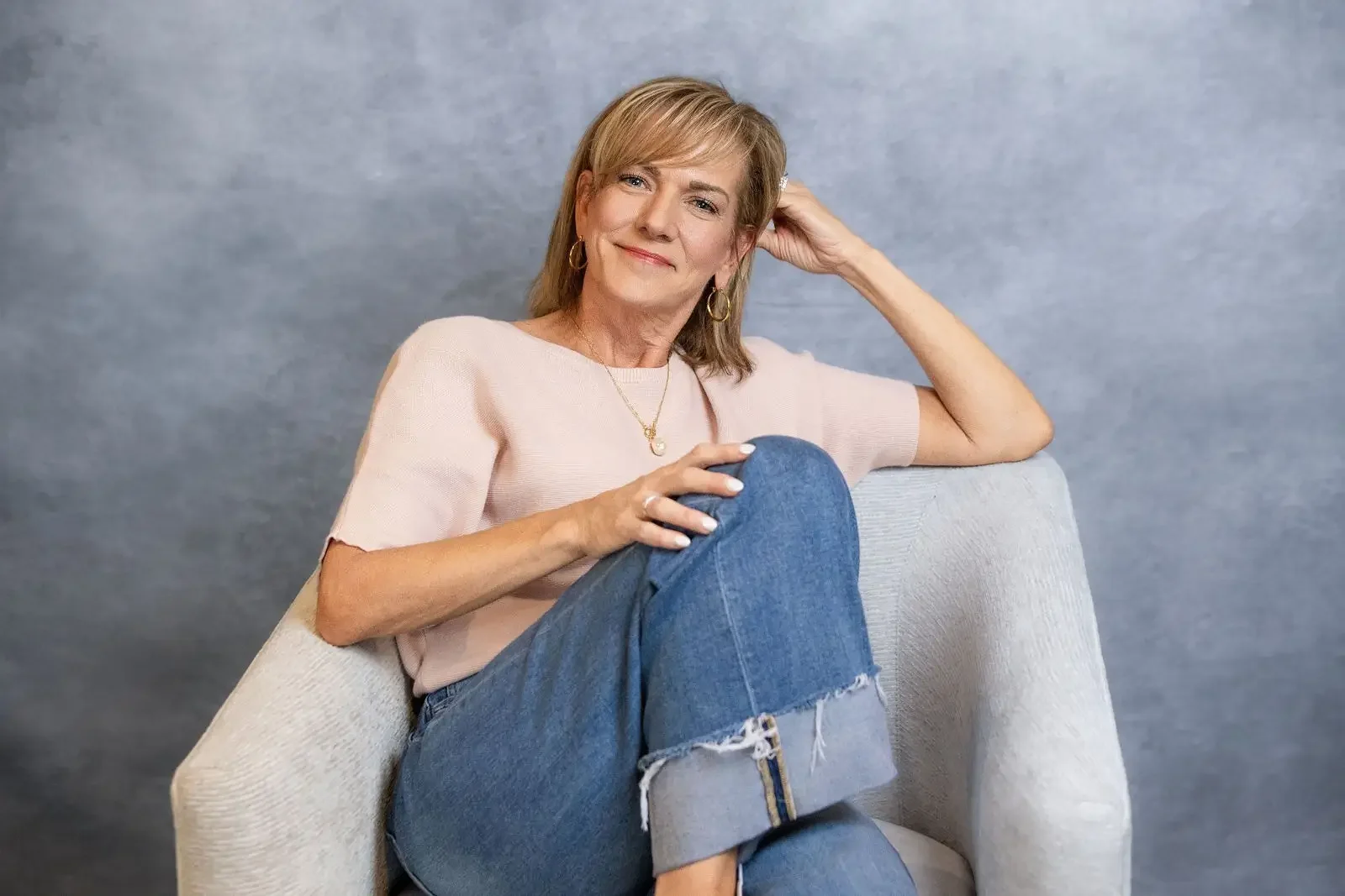 A middle-aged woman with blonde hair, wearing a pink shirt and blue jeans, sitting comfortably on a light-colored armchair against a neutral gray background. She is smiling gently, with her left hand resting behind her head and her right hand on her knee.