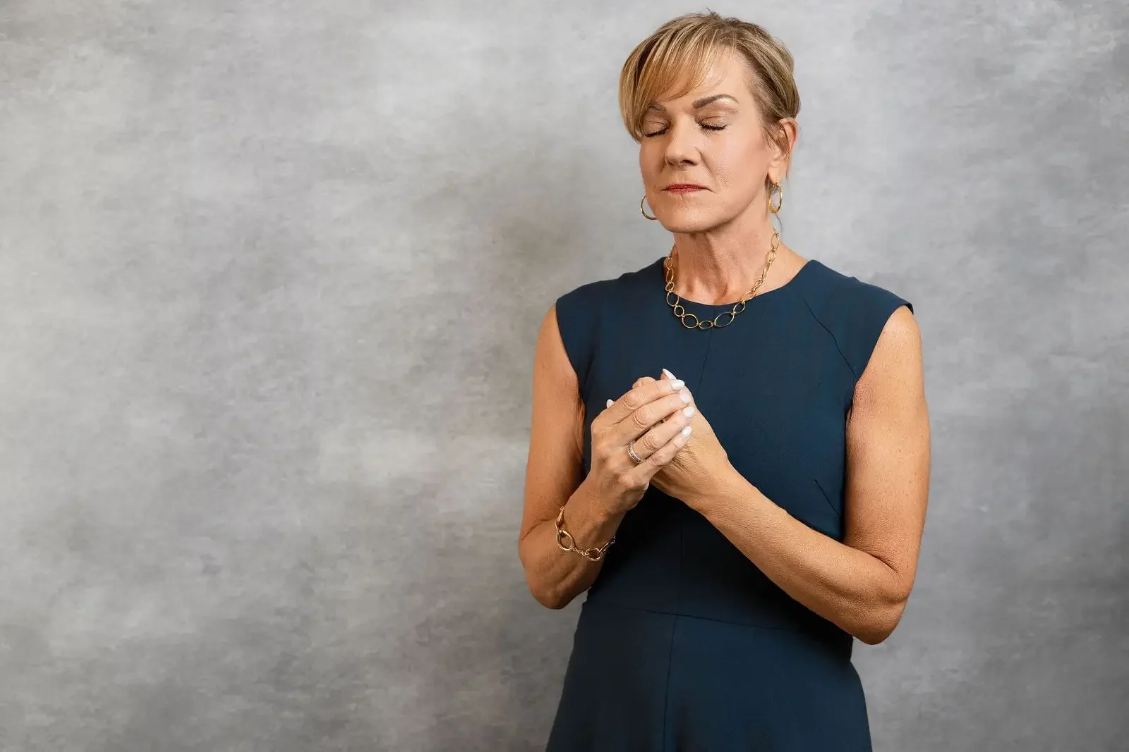 Darlene Lekowski, author, speaker, and survivor advocate in a navy sleeveless dress with her eyes closed, holding her hands close to her chest, standing against a gray background.