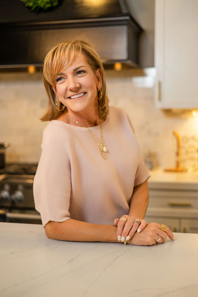Darlene Lekowski, author, speaker, and survivor advocate with short, blond hair wearing gold hoop earrings and layered jewelry, standing in a modern kitchen with a white countertop, beige cabinets, and a black range hood.
