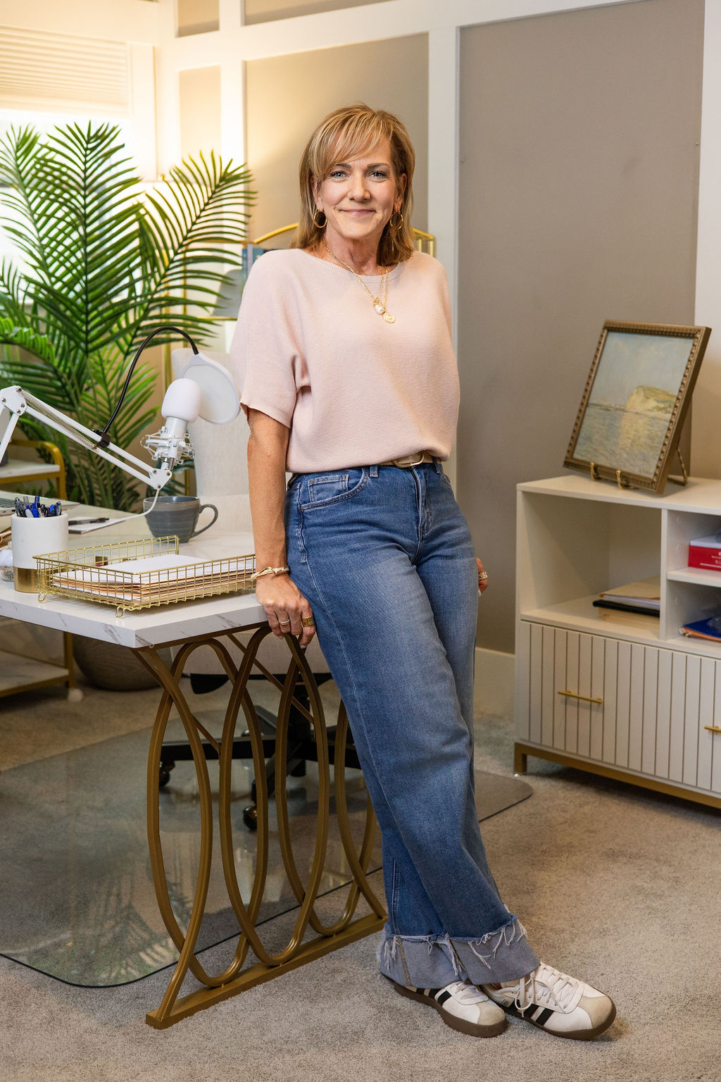 A woman standing in an office space with a smile, wearing a light pink top, blue jeans, and sneakers, with office supplies and a potted plant in the background.