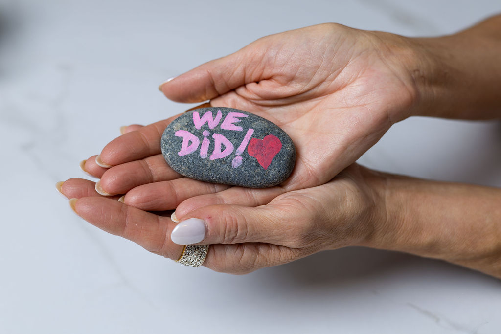 A pair of hands holding a small, oval stone with the words "WE DID!" written in pink and a red heart painted on it.