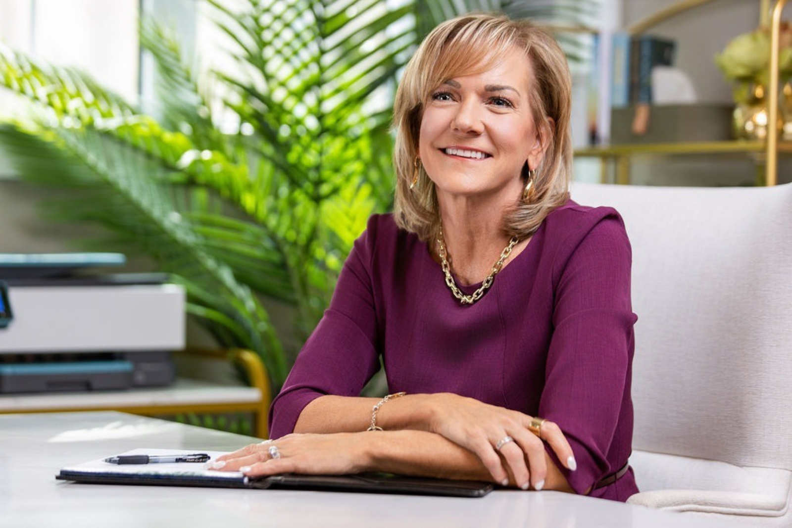 A smiling woman with shoulder-length blonde hair wearing a purple dress, gold jewelry, sitting at a desk with a notepad, pen, and a printer, surrounded by green plants in a well-lit office.
