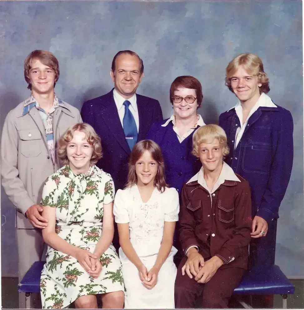 Group portrait of seven people, five young adults and two middle-aged adults, posing against a studio backdrop. The image appears to be from the 1970s, based on their clothing styles. Four are sitting on a bench, and three are standing behind them.