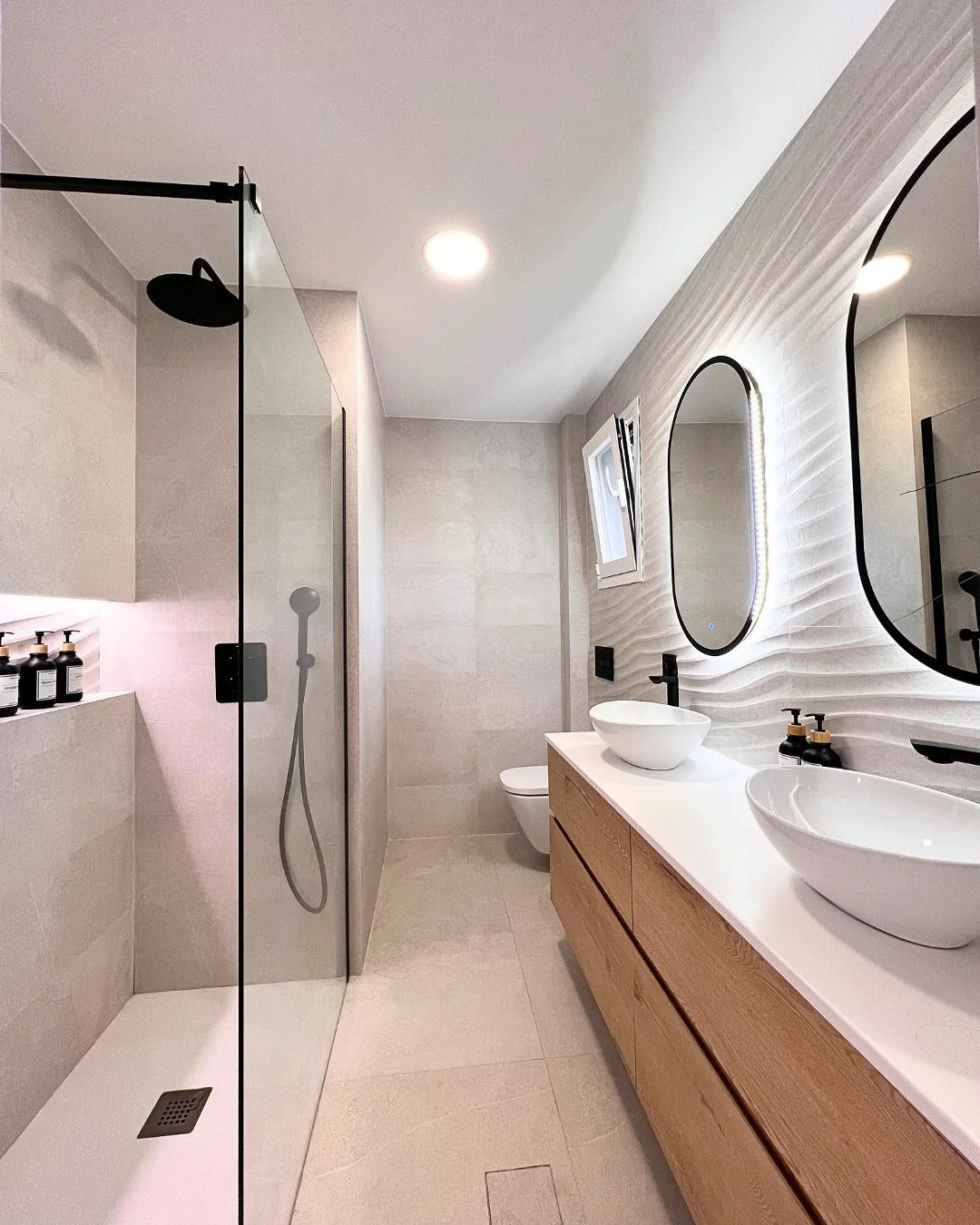 Modern bathroom with walk-in shower, double sinks with vessel basins, a large mirror with backlit lighting, beige tiled walls and floor, a small window with natural light, and minimalist fixtures.