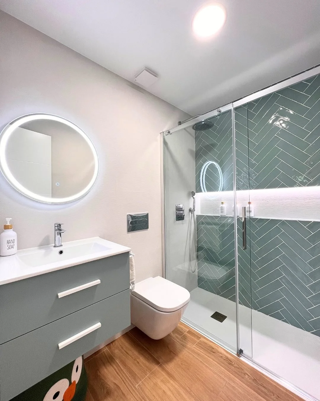 Modern bathroom with a circular illuminated mirror above a white vanity with a hand soap dispenser, a wall-mounted toilet, and a glass shower enclosure with green herringbone tiles and built-in shelves.