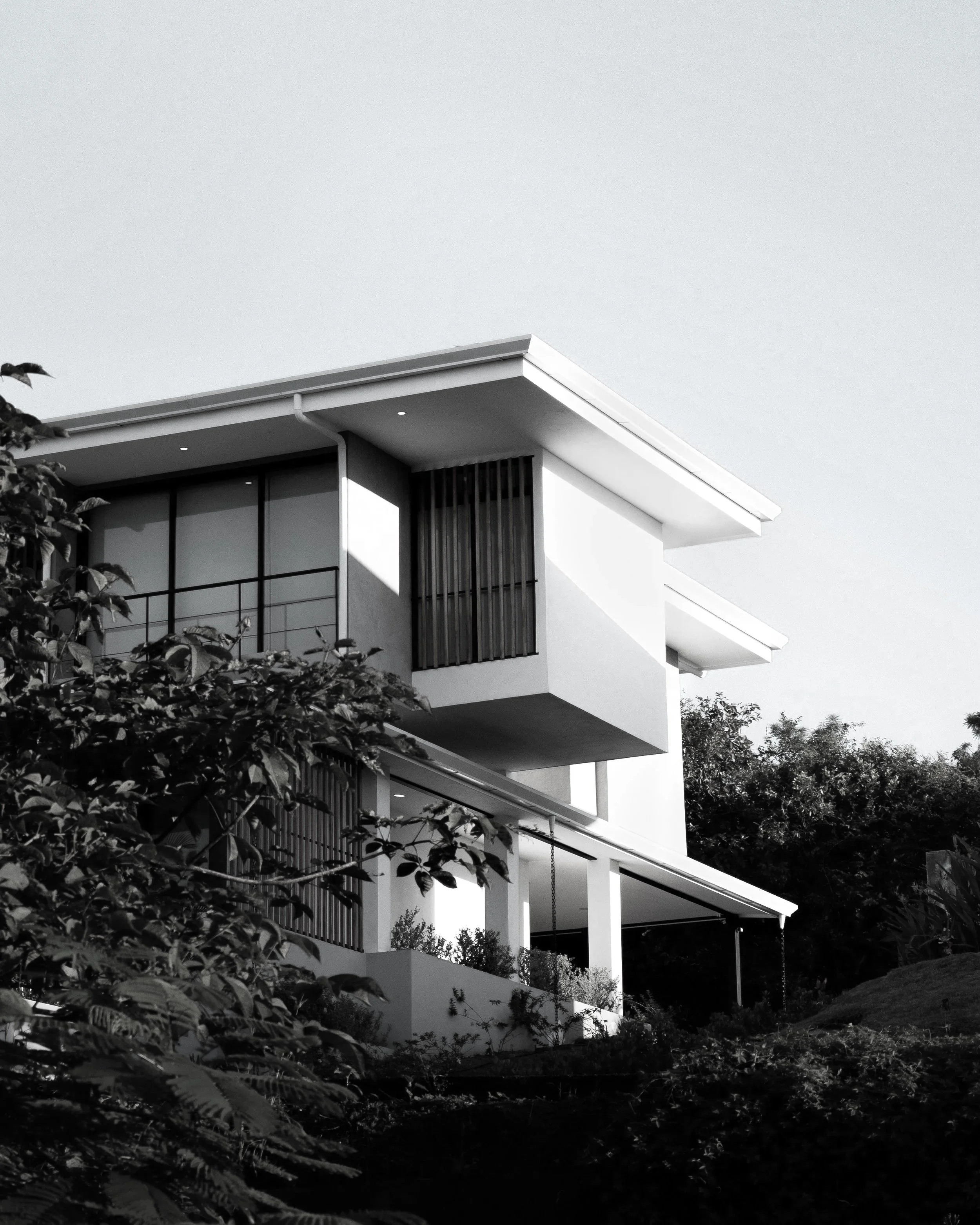 Modern white multi-story house with large glass windows and balconies, surrounded by trees and plants, under a clear sky.