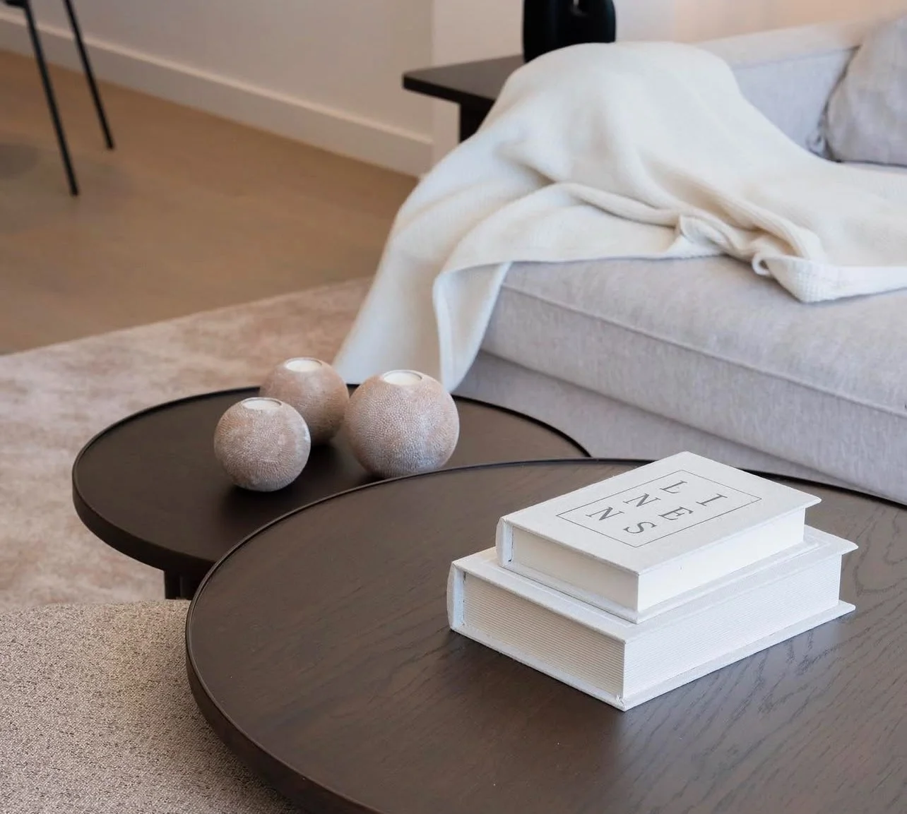 Two dark round coffee tables with three beige textured spherical candle holders, two stacked white books with black text, and a light-colored sofa with a white blanket in a modern living room.