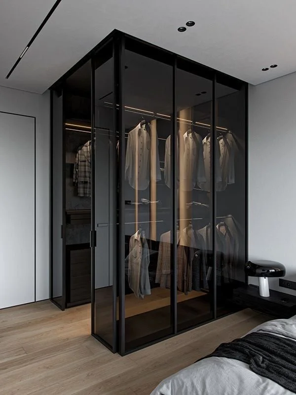 Modern bedroom with a black glass wardrobe with sliding doors, containing neatly hung clothes. A bed with white and black linens is partially visible in the foreground, and a small speaker or device is on a black side table beside the bed.