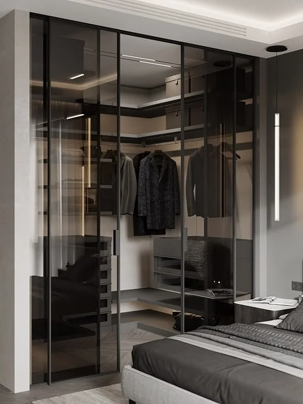 Modern bedroom with a glass-walled closet containing hanging clothes, a bed with black and gray bedding, and a small sitting area with a chair and side table.
