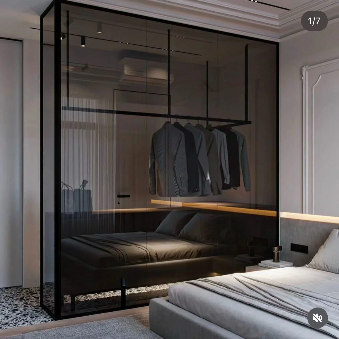 Modern bedroom with a glass partition and hanging clothes, featuring a bed with gray bedding, nightstand, and decorative interior design.