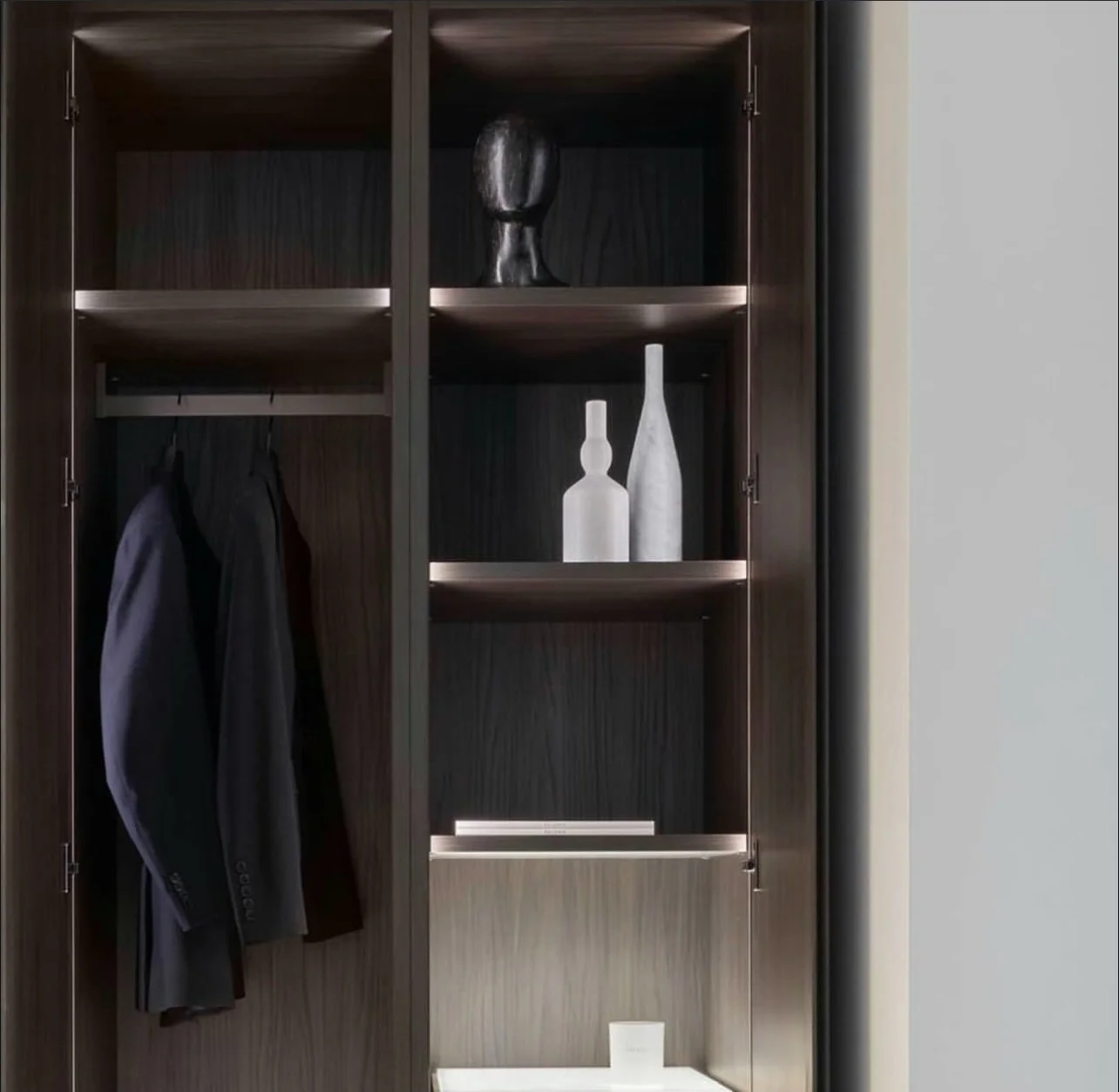 Open wooden closet with two suits hanging inside, black decorative vase on top shelf, two white decorative bottles on middle shelf, white candle on bottom shelf.