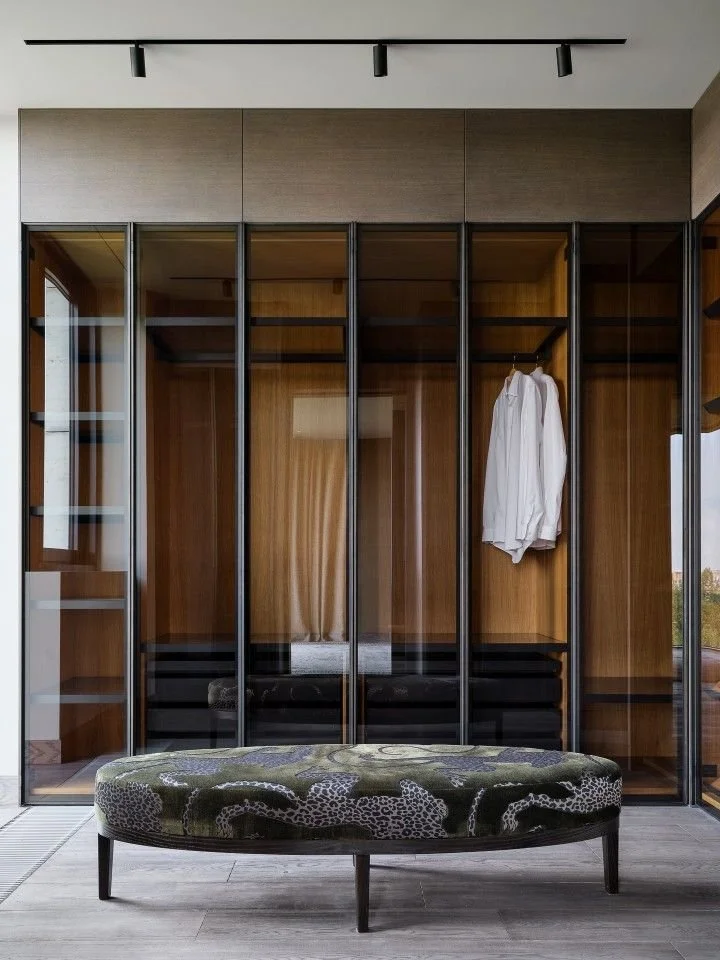 Modern glass-walled wardrobe with wooden interior and a white shirt hanging inside, with a padded bench with animal print fabric in the foreground.