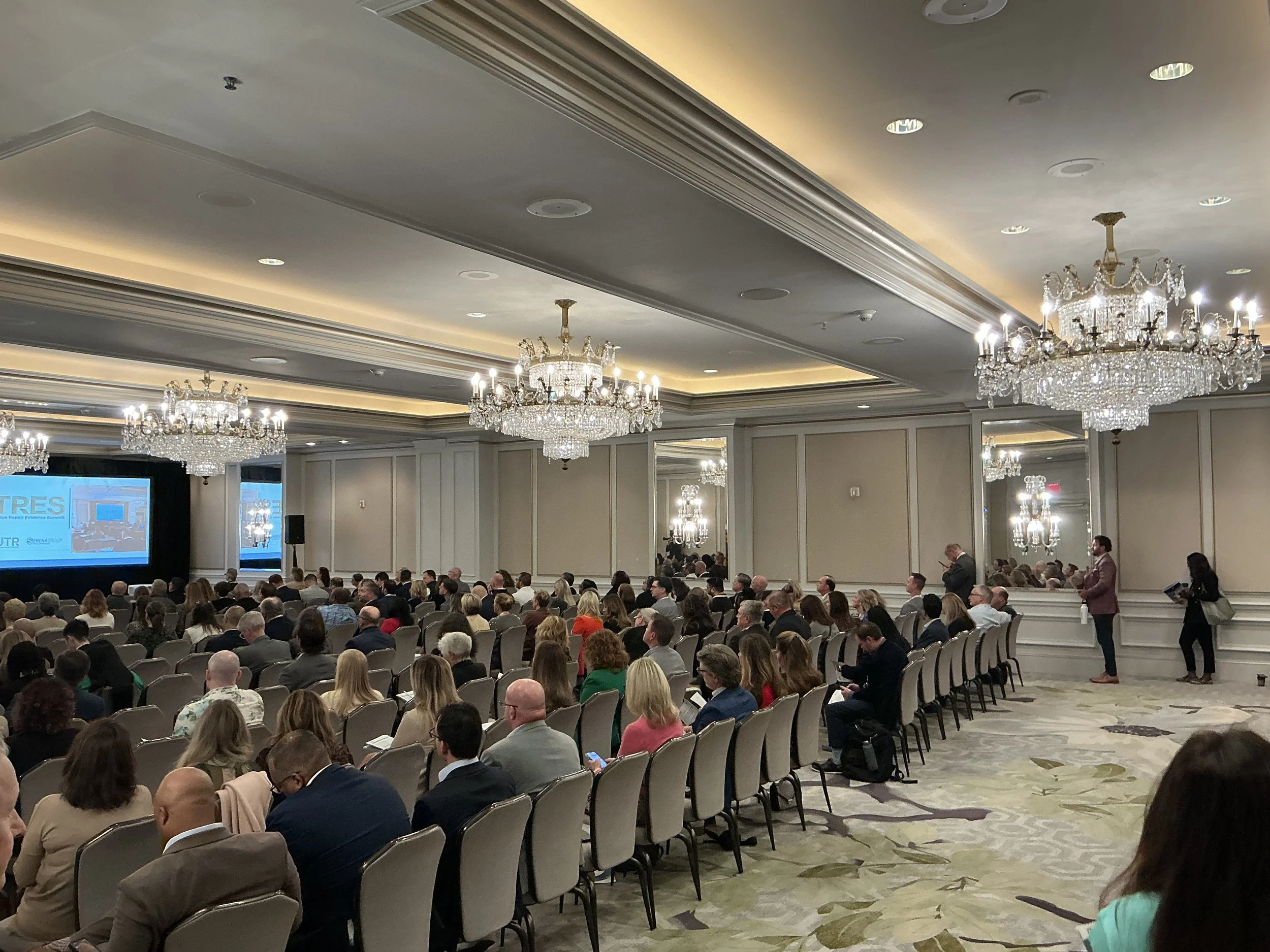 Large conference room with chandeliers and many seated attendees facing a stage with a large screen.