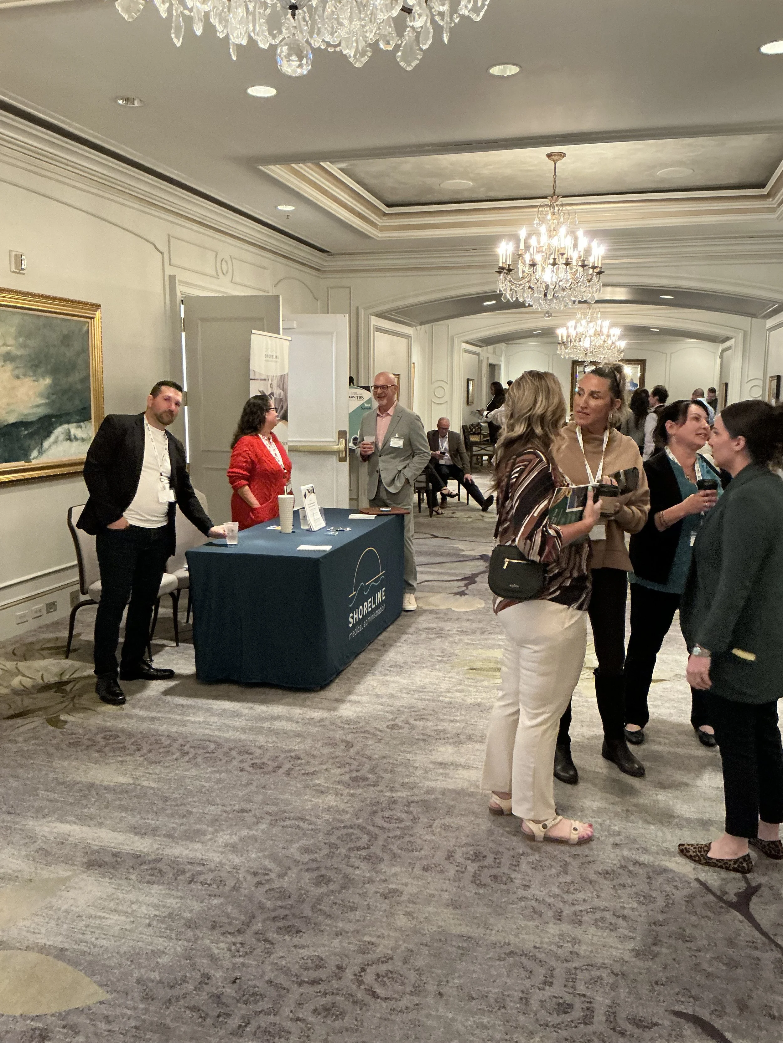 People networking at a conference or business event in a well-lit room with chandeliers, a Shoreline display table, and framed artwork on the walls.