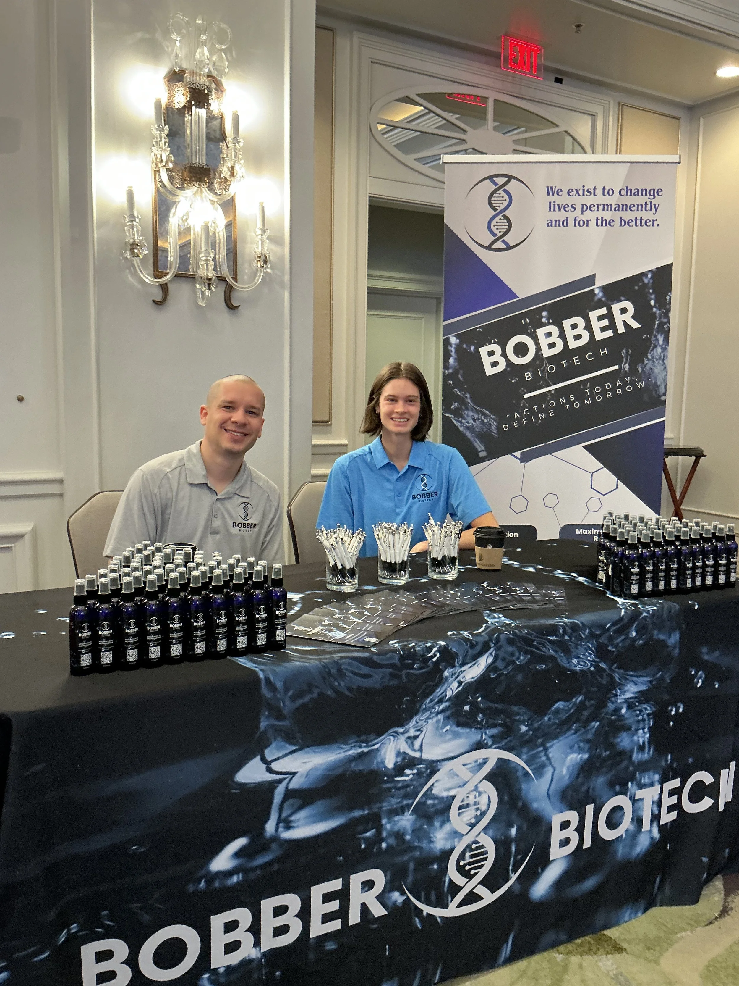 Two smiling people sitting behind a promotional table for Bobber Biotech, with bottles and pens on the table, in a room with ornate wall decor and a large banner displaying the company's logo and slogan nearby.