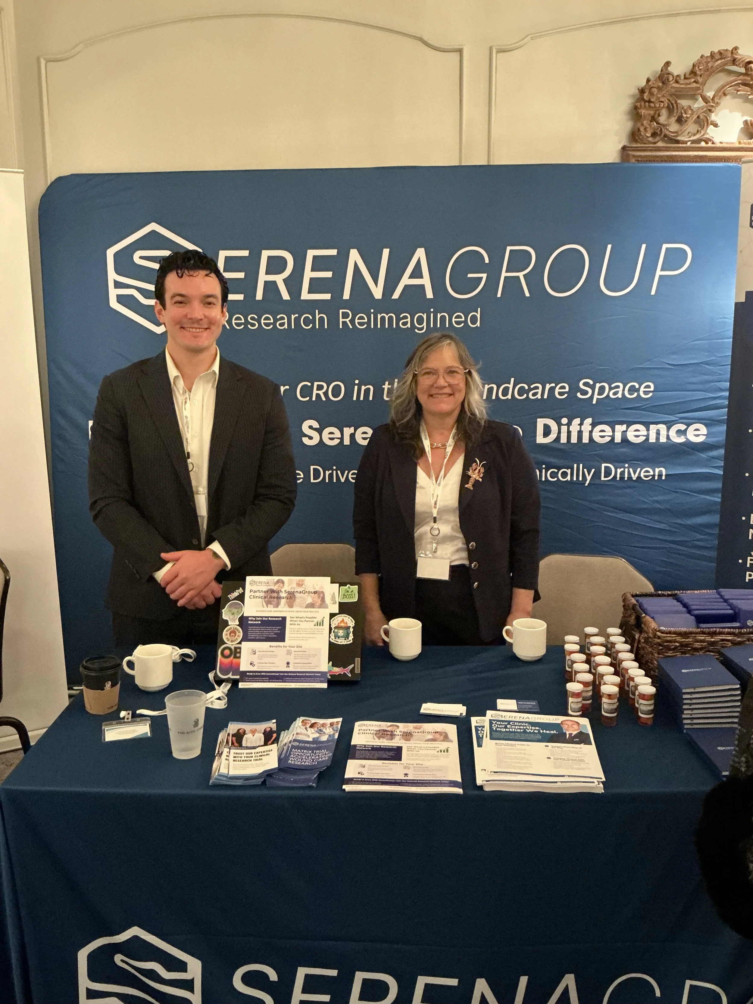 Two people standing behind a table at a conference booth for Serena Group, smiling. The table has brochures, buttons, and coffee mugs. The backdrop displays the Serena Group logo and slogan.