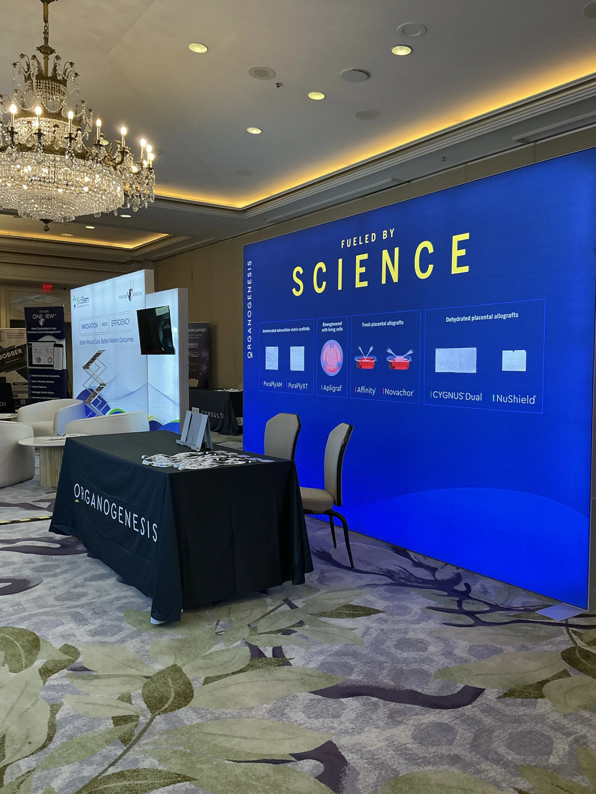 Exhibit booth with a large blue digital screen displaying information about science, organization name 'Organogenesis' on a black tablecloth, chairs, chandelier, and carpeting with leaf pattern in a conference room.