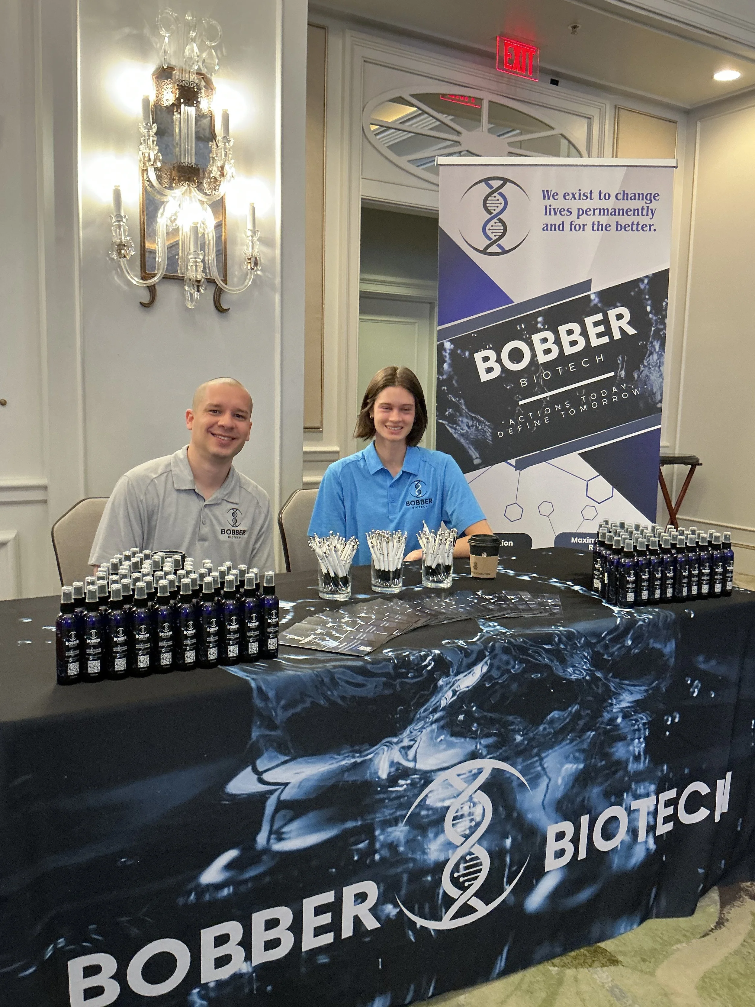Two people, a man and a woman, sitting at a booth for Bobber Biotech at an event. They are smiling and wearing shirts with the company logo. The booth has a table covered with a black tablecloth featuring the Bobber Biotech logo, with bottled product