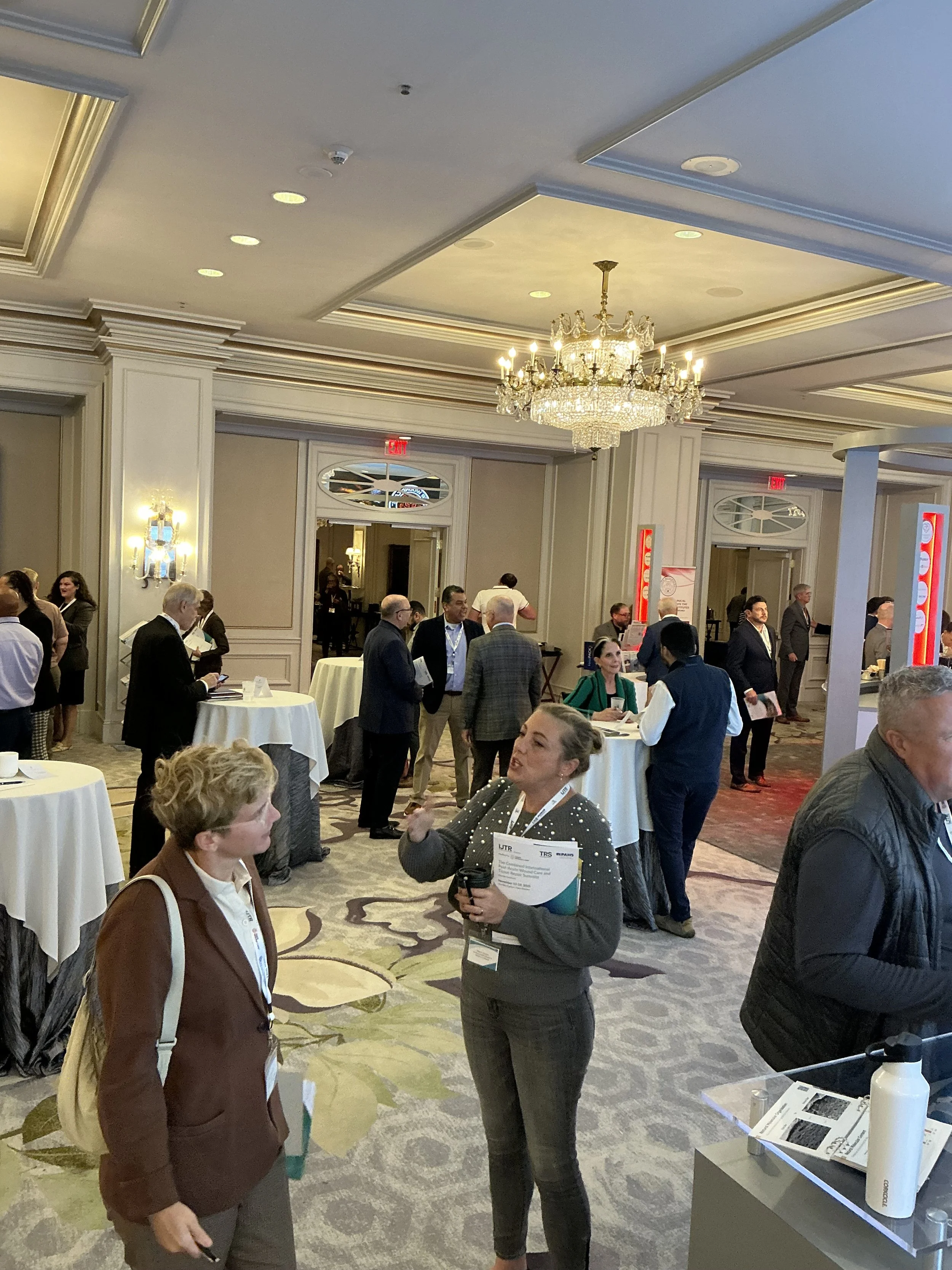 People socializing and networking at a conference or event in an elegant hotel ballroom with chandeliers and round tables.