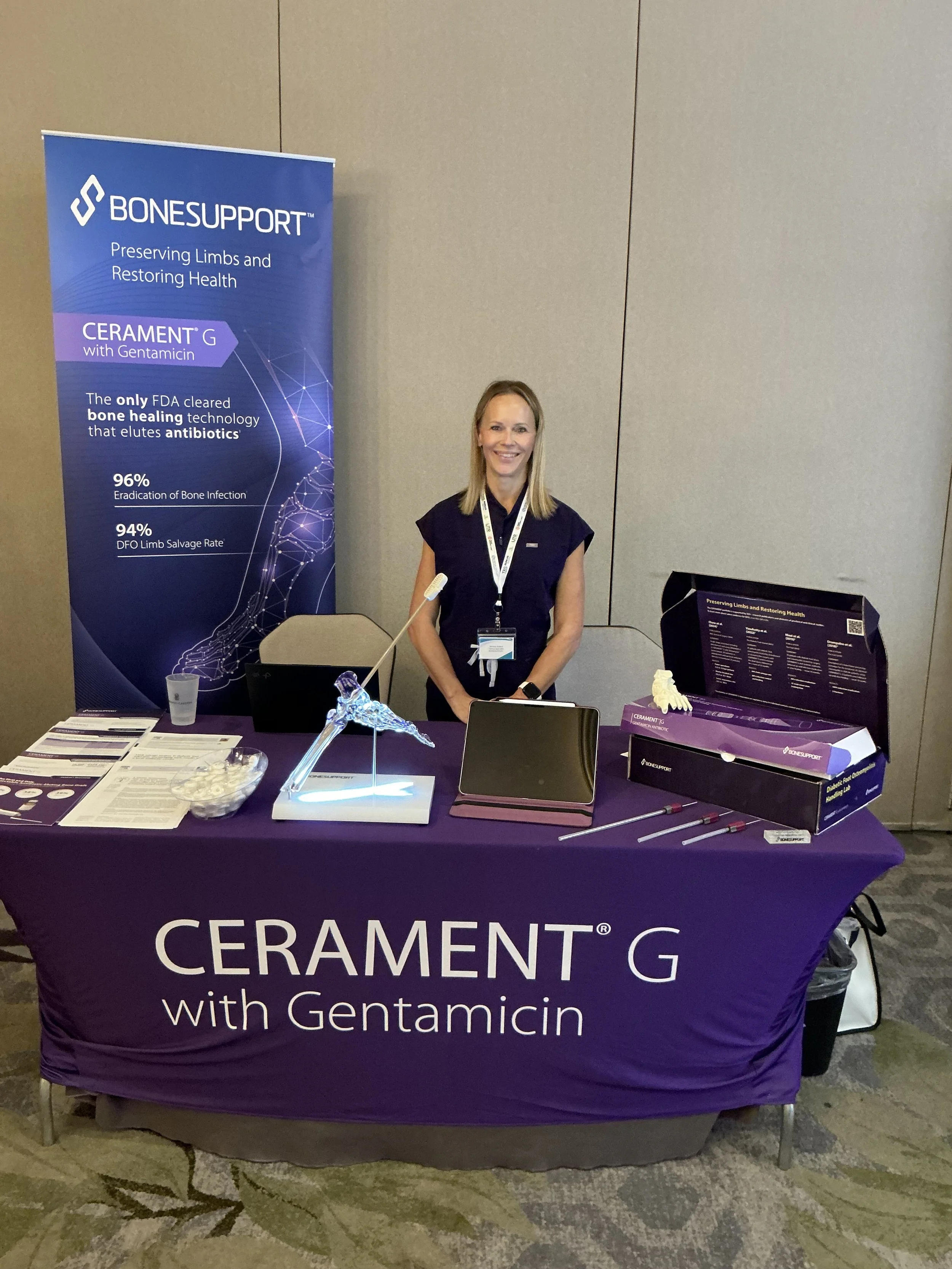 A woman standing behind a table at a conference booth for BONESUPPORT. The table is covered with a purple cloth displaying the words 'CERAMENT G with Gentamicin'. The booth has a large vertical banner promoting Bone Support's technology for preservin