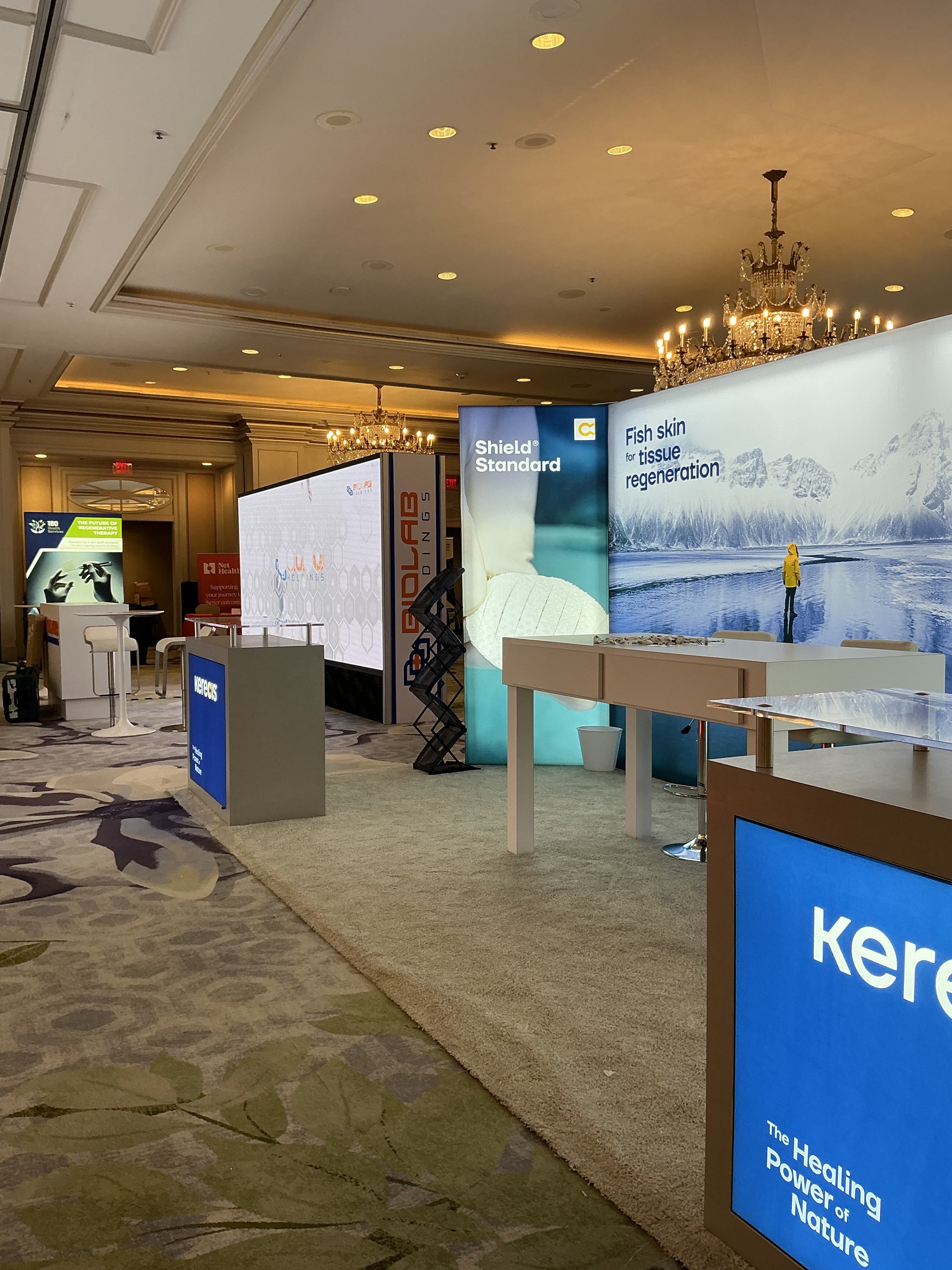 Exhibition booth showcasing skincare products with digital displays, large landscape images, and product information, set in an elegant ballroom with chandeliers and decorative carpeting.