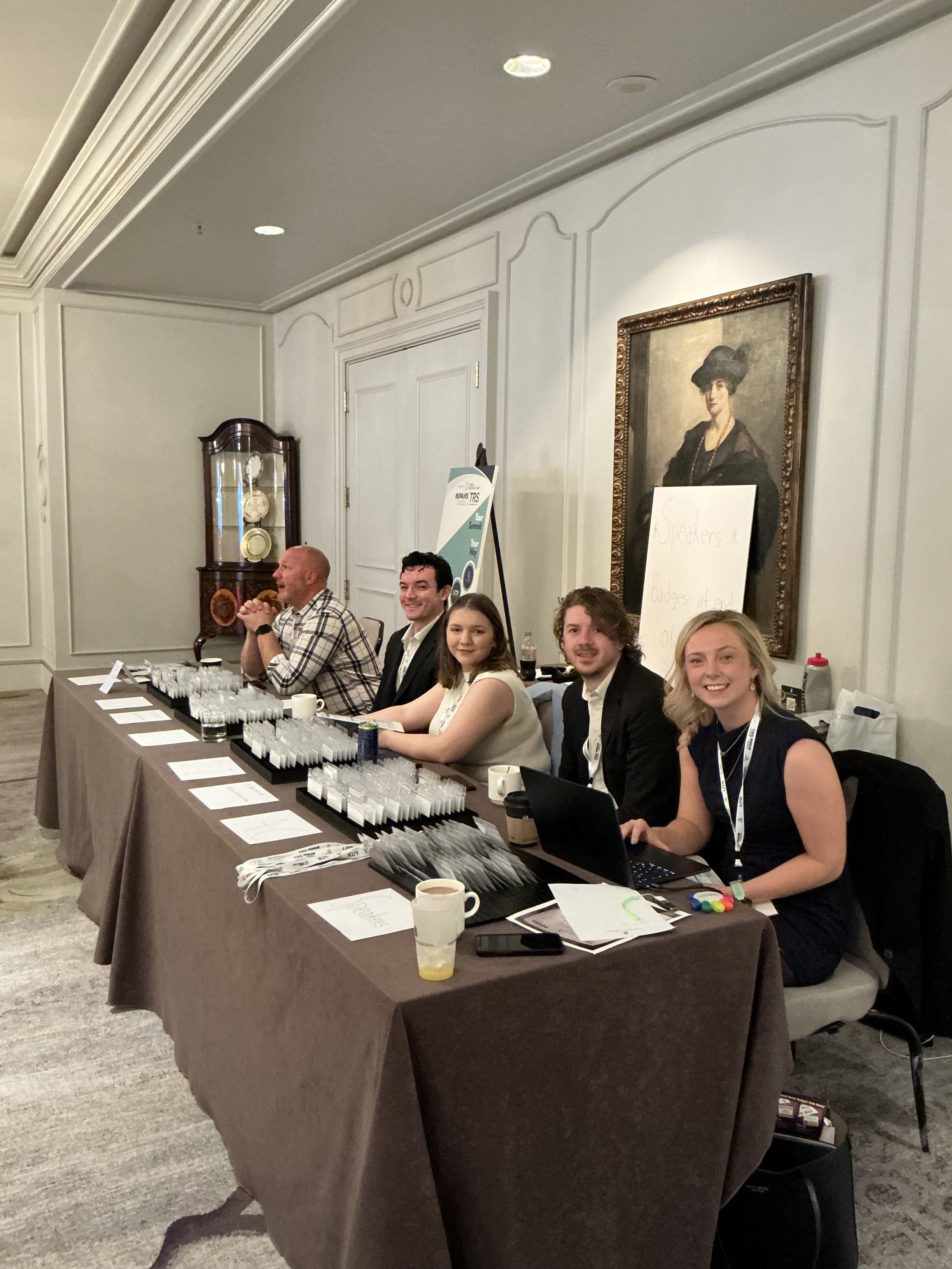 Six people sitting at a registration or check-in table at an event, with papers, pens, and glasses arranged on the table in front of them.