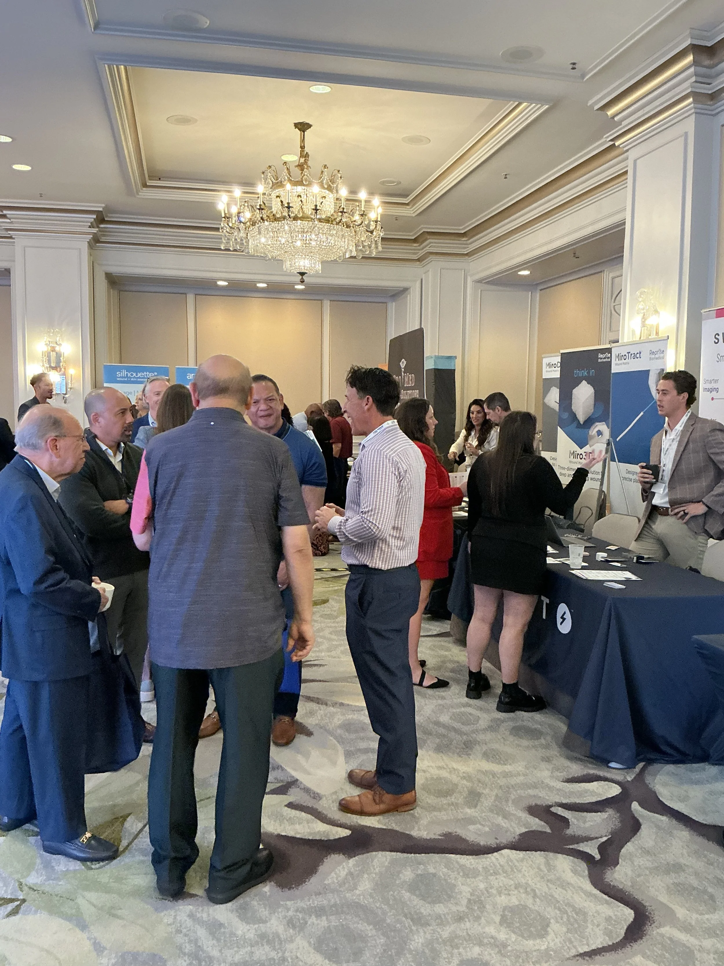 People at a conference, engaging in conversation near exhibitor booths, in an elegant room with a chandelier, crown molding, and patterned carpet.