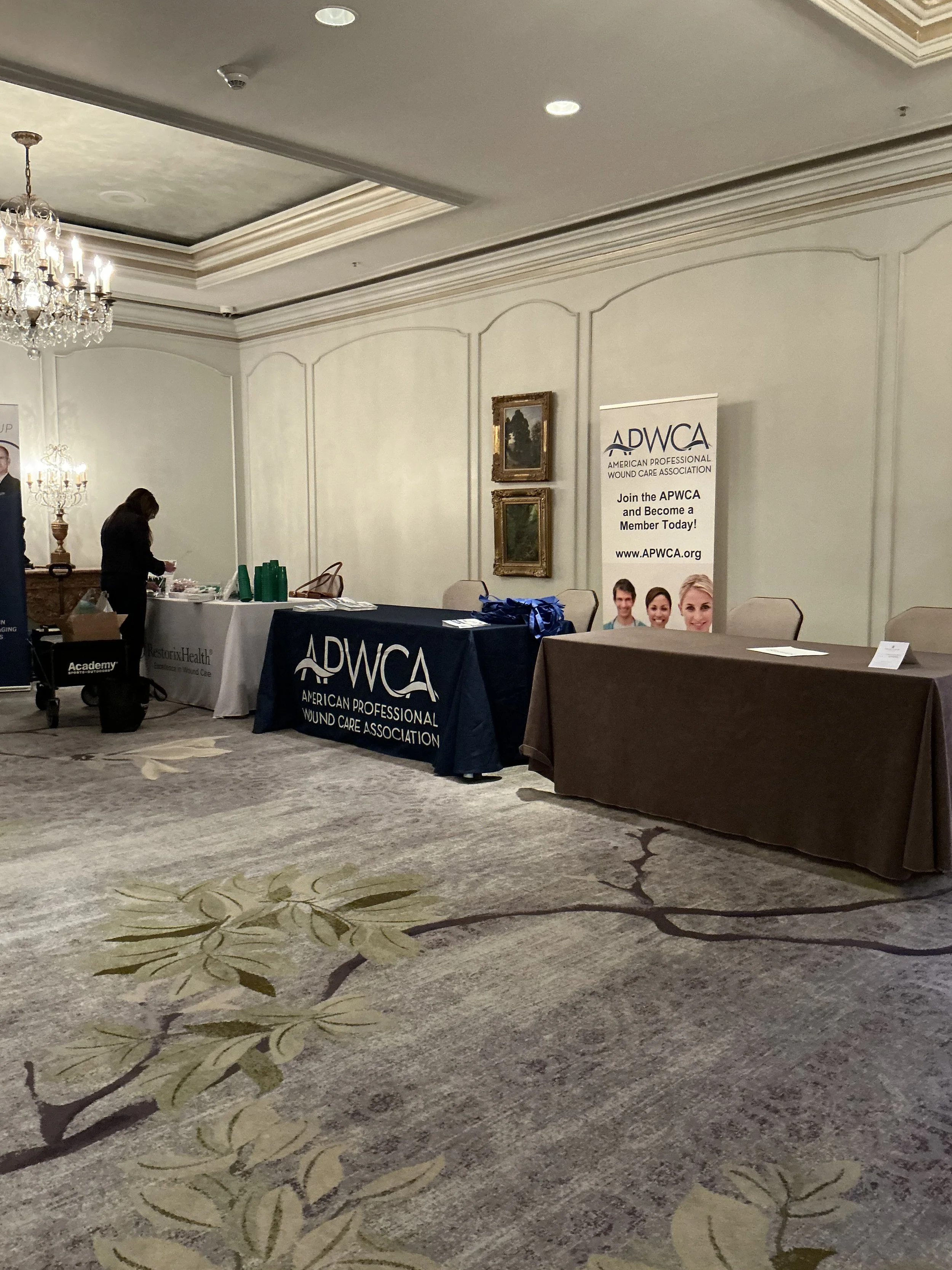 Conference room with tables displaying banners for the American Professional Wound Care Association (APWCA) and RestorixHealth, with a woman setting up materials and a chandelier hanging from the ceiling.