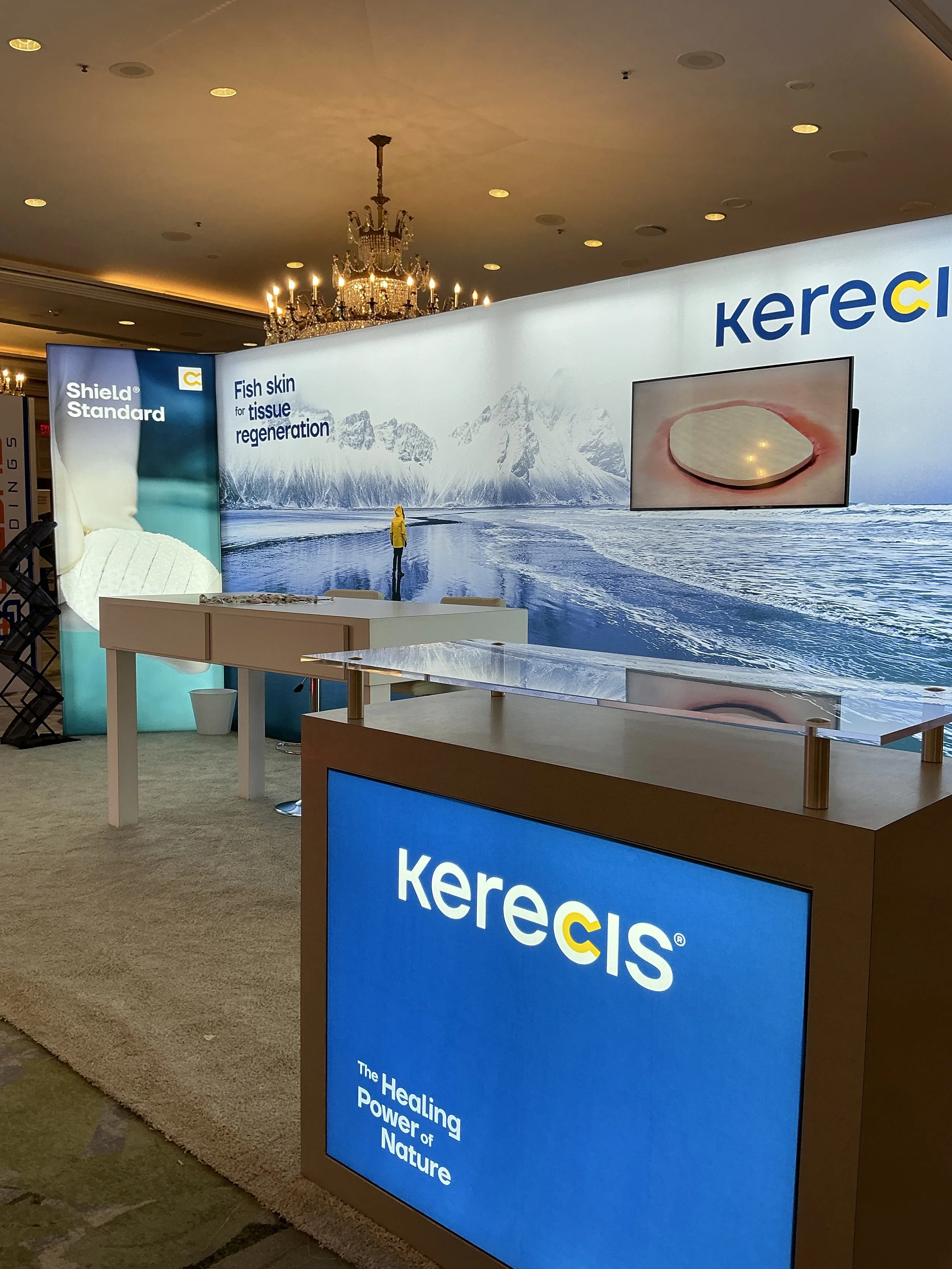 Trade show booth for Kerecis featuring a large banner with icy mountain scenery and the slogan 'Fish skin for tissue regeneration', a lit chandelier in the ceiling, a display with the Kerecis logo and the words 'The Healing Power of Nature.'