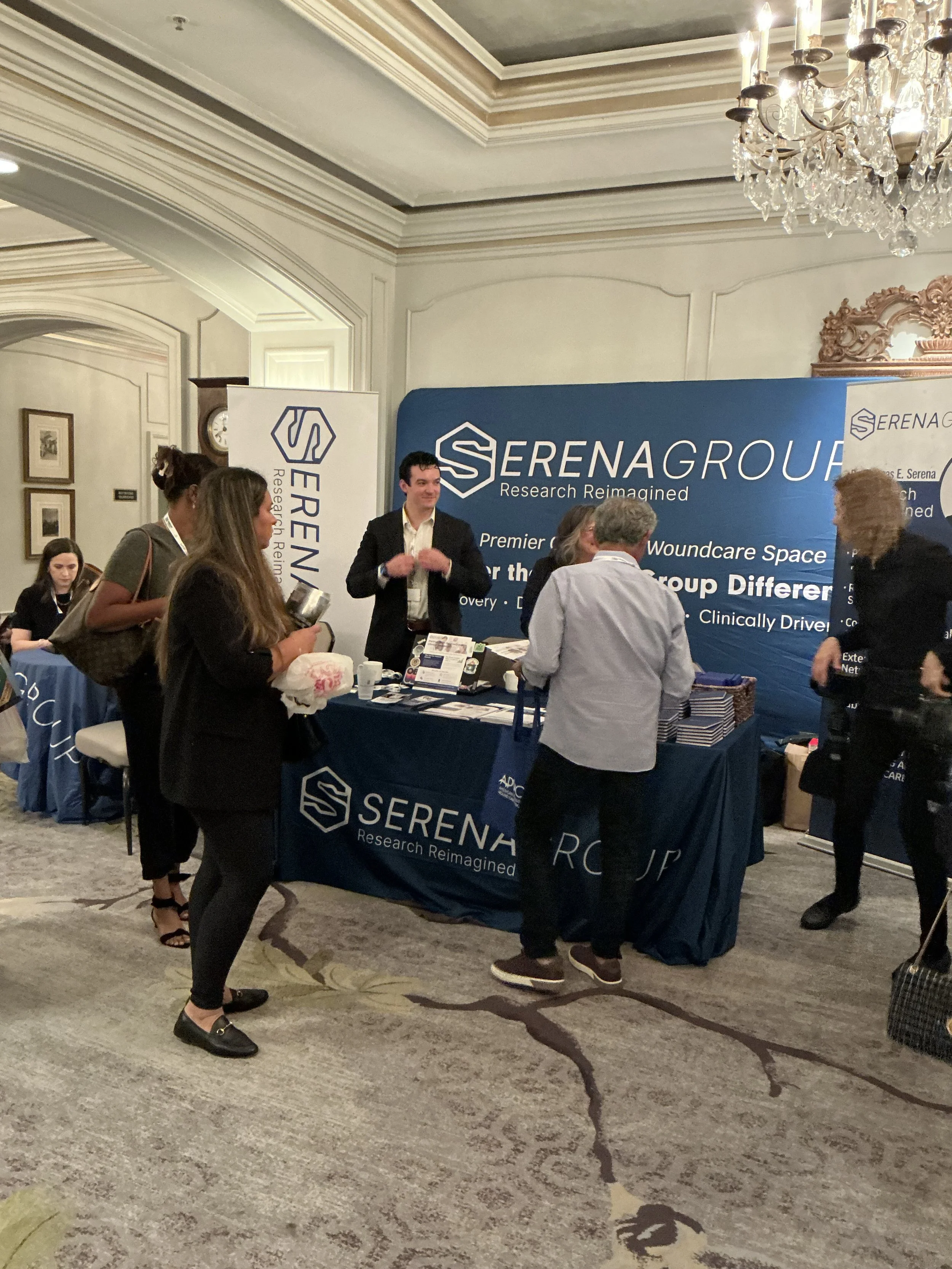 People gathered around a booth at a conference or event, with a large blue banner displaying 'SERENAGROUP - Research Reimagined' and a table covered with promotional materials.