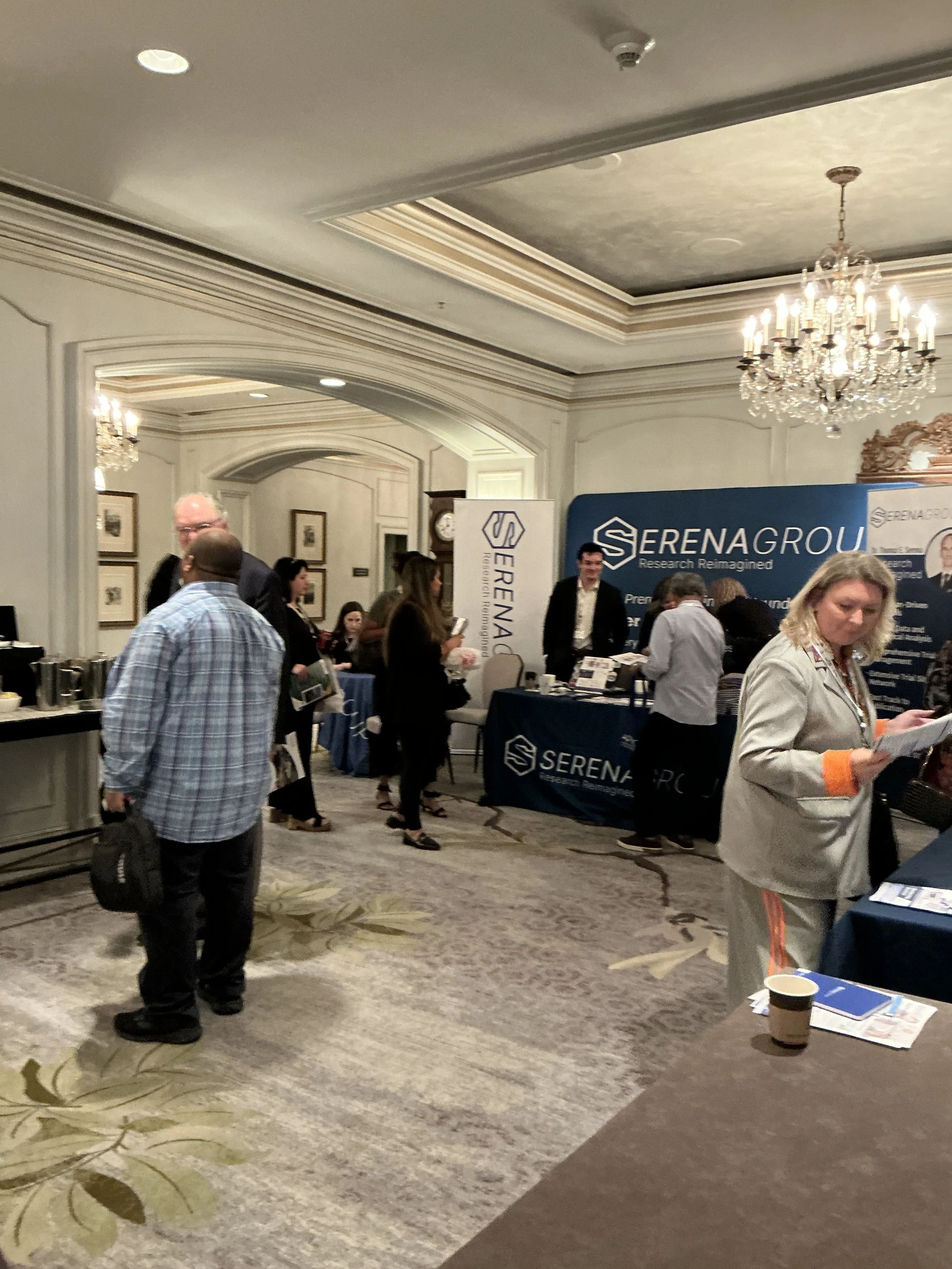 People attending a conference in a luxurious banquet hall, with a Serena Group booth set up, decorated with banners and informational materials.